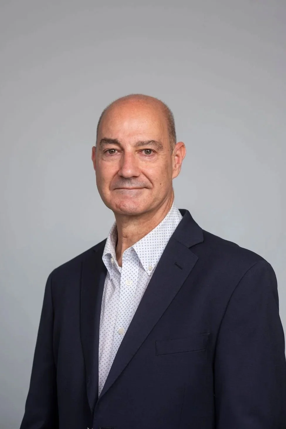 A middle-aged man with a shaved head in a business suit, standing against a plain, light gray background.