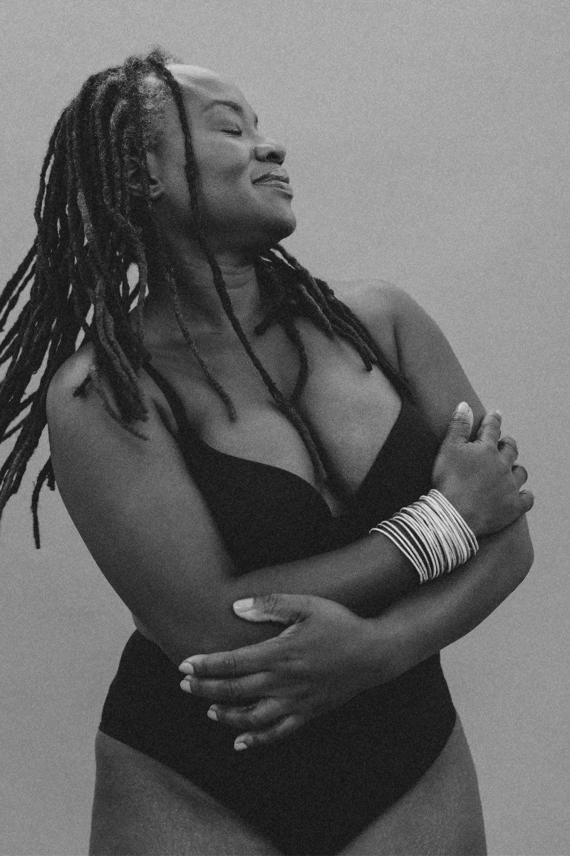 Black and white portrait of a woman with dreadlocks, wearing a black top and multiple bangles, standing against a plain background with her arms crossed, eyes closed, and a gentle smile.