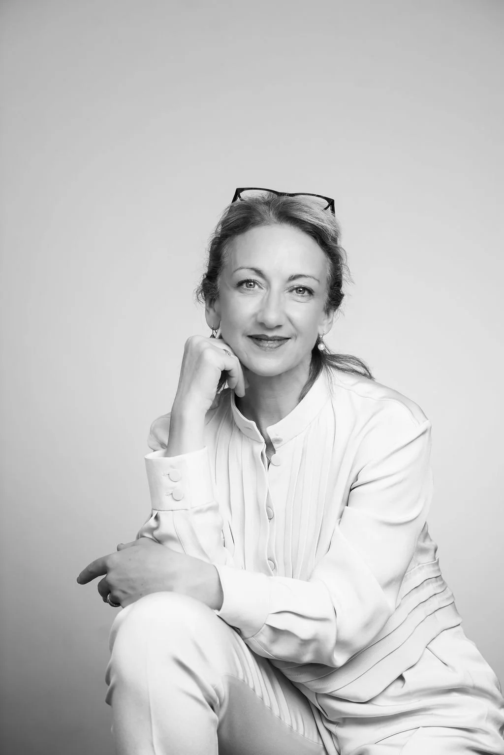 A black and white portrait of a woman with glasses on her head, sitting with her chin resting on her hand, smiling softly at the camera.