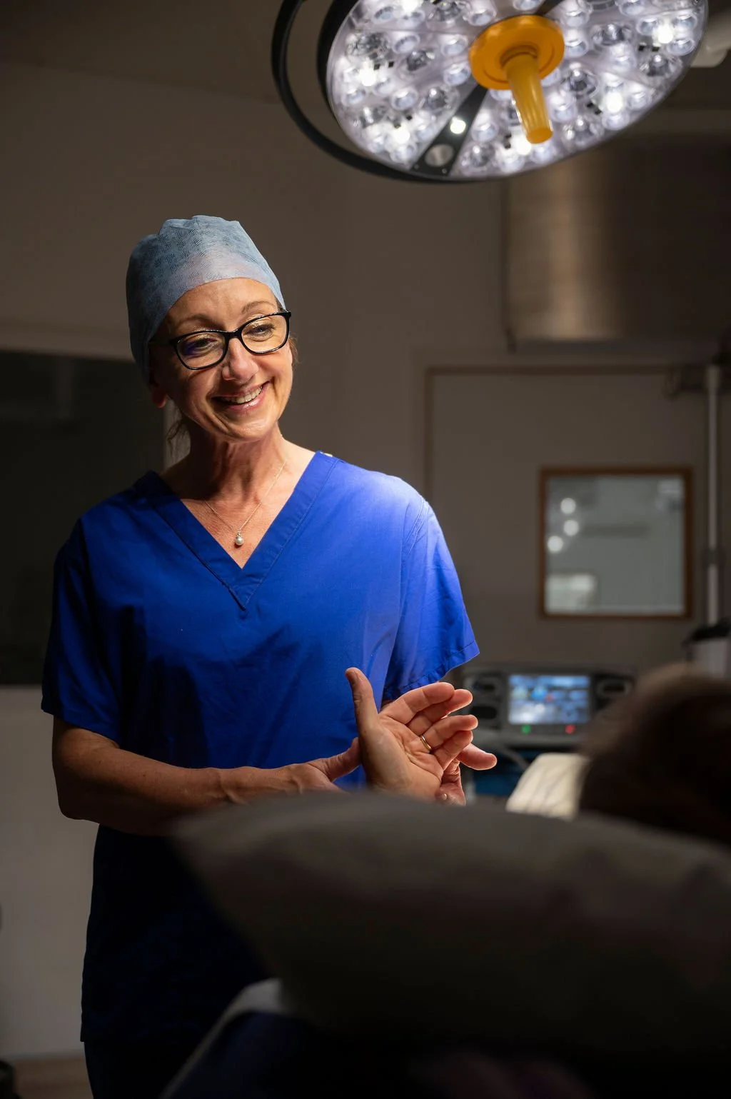 A smiling female healthcare professional in blue scrubs and a surgical cap talking to a patient in an operating room.
