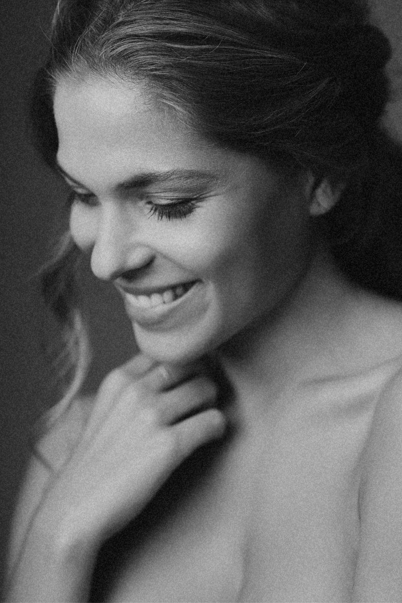 A black and white close-up portrait of a woman smiling with her eyes closed, touching her chin with her hand, hair styled back.