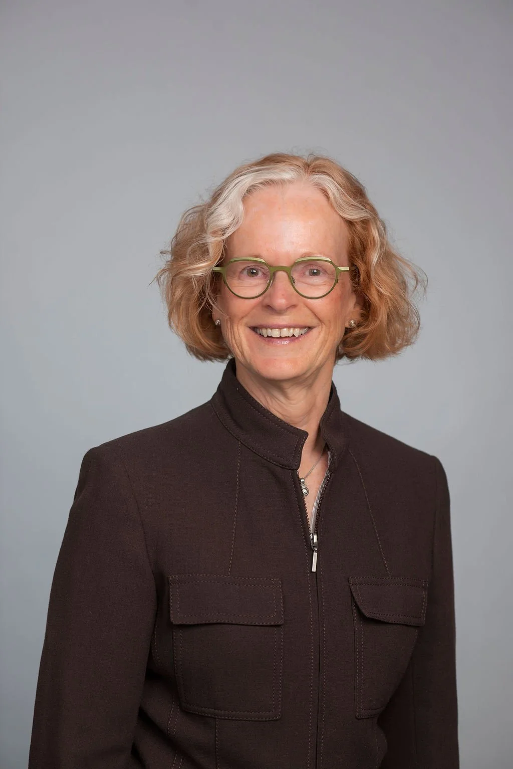 A smiling woman with short, curly blonde hair wearing green glasses and a dark zip-up jacket against a neutral background.