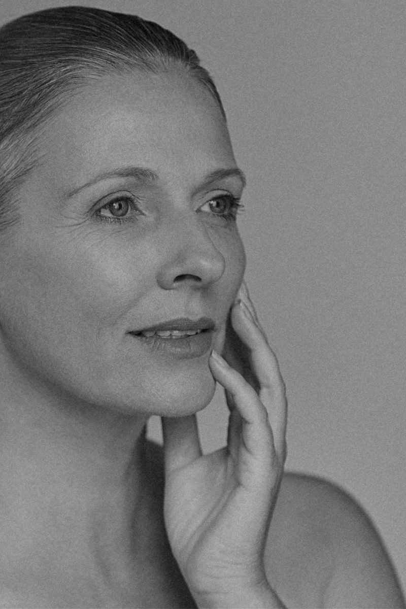 Black and white portrait of a woman with light-colored eyes, slicked-back hair, and light makeup, touching her face.