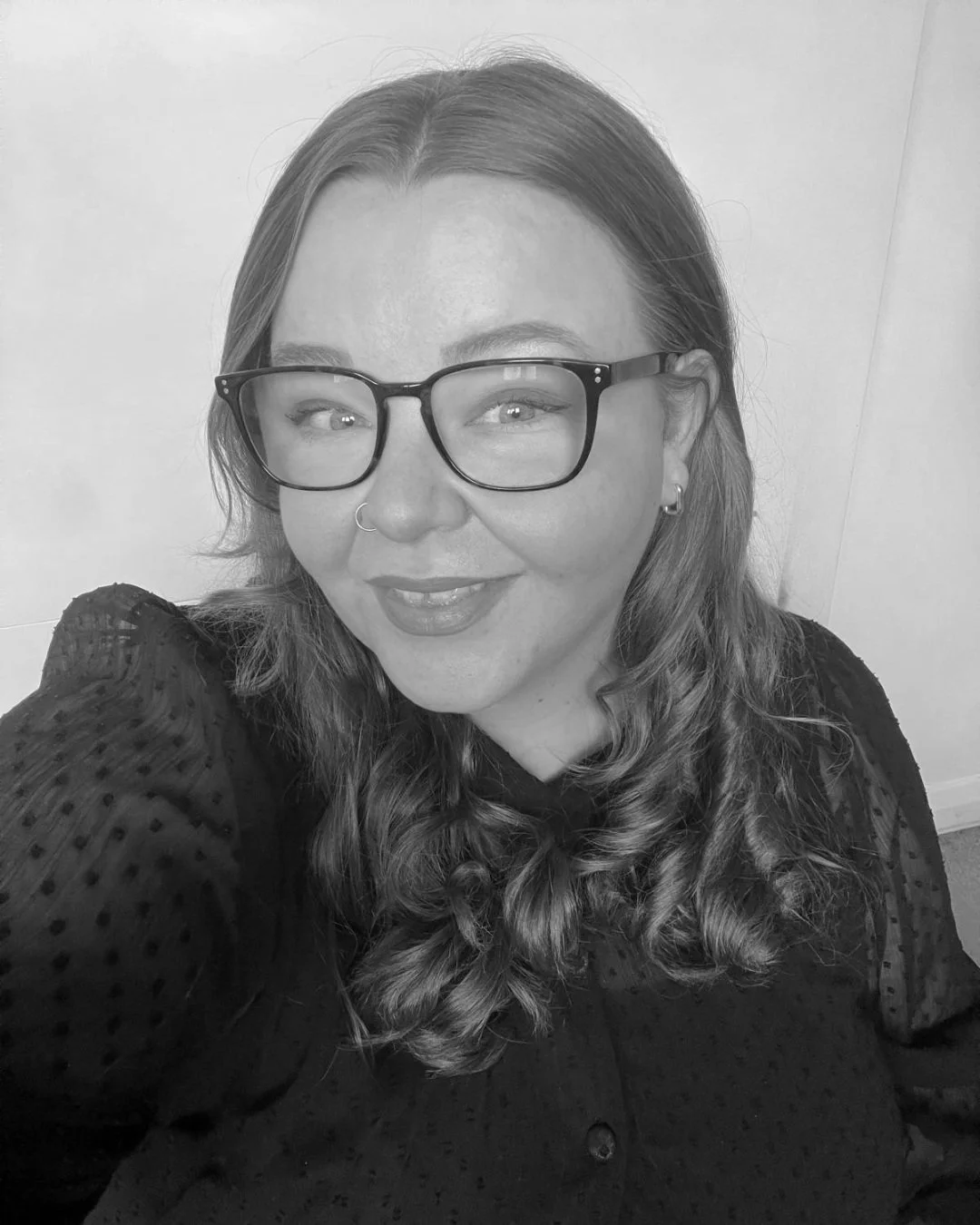 A woman with long wavy hair wearing glasses, a nose ring, and earrings, smiling at the camera in a black shirt with textured dots.