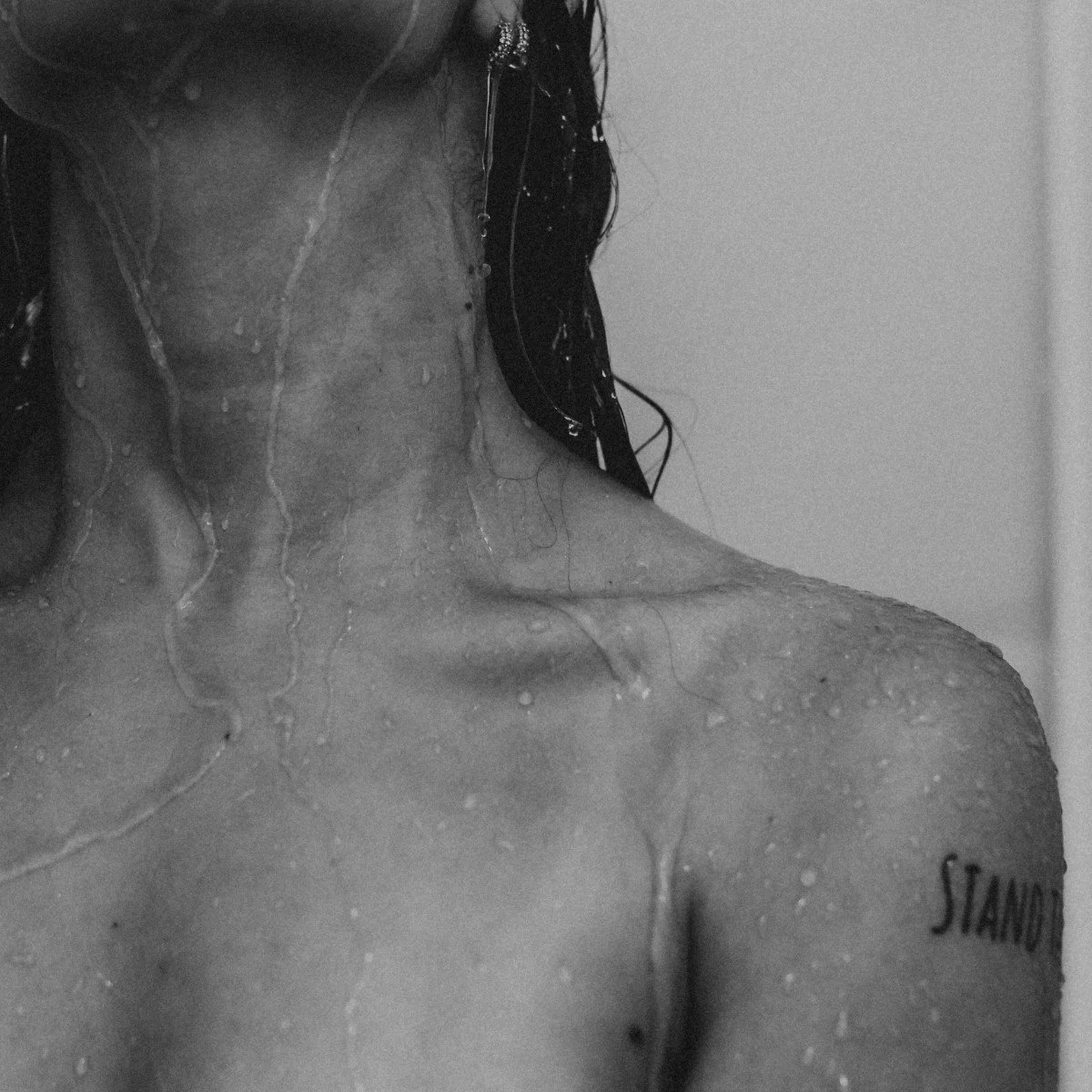 Close-up black and white photo of a wet female shoulder with a tattoo that reads 'STAND' and dripping water.