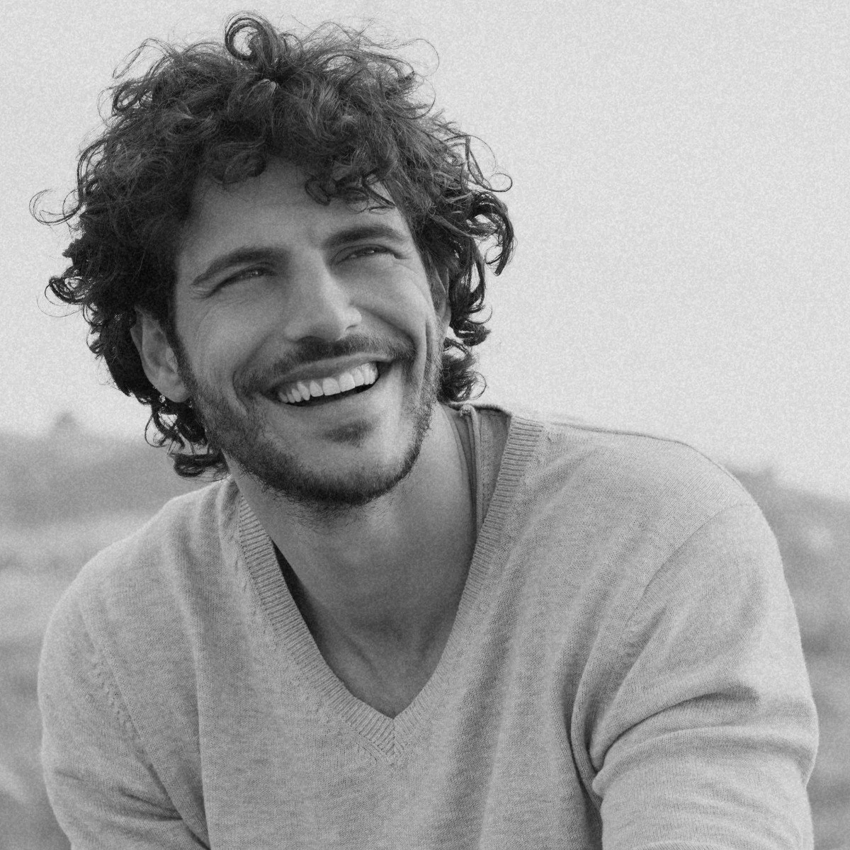 Black and white photo of a man with curly hair and a beard, smiling with his teeth showing, wearing a light sweater outdoors.