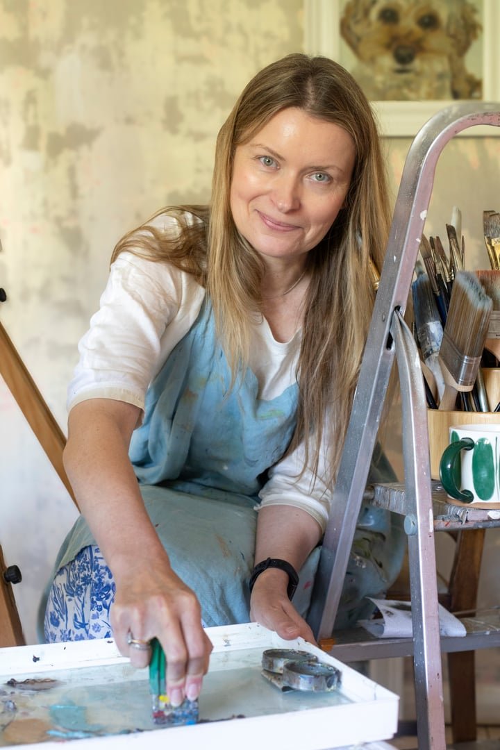 A woman with long blonde hair wearing a white shirt and blue apron, smiling, engaged in painting or mixing colors in an artist's studio. There is a ladder with brushes and a cabinet with a mug and art supplies. A framed portrait of a dog is on the wall in the background.