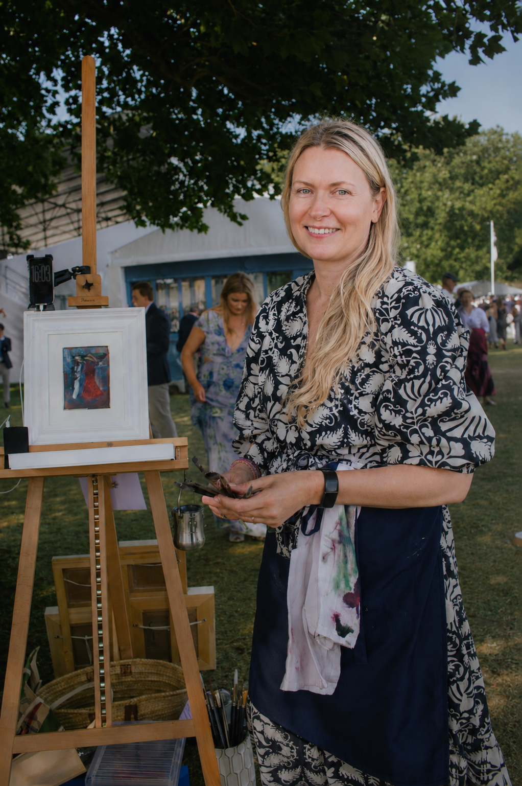 Woman with blonde hair in an outdoor art market, standing next to an easel with framed artwork, wearing a floral black and white blazer and a blue apron.