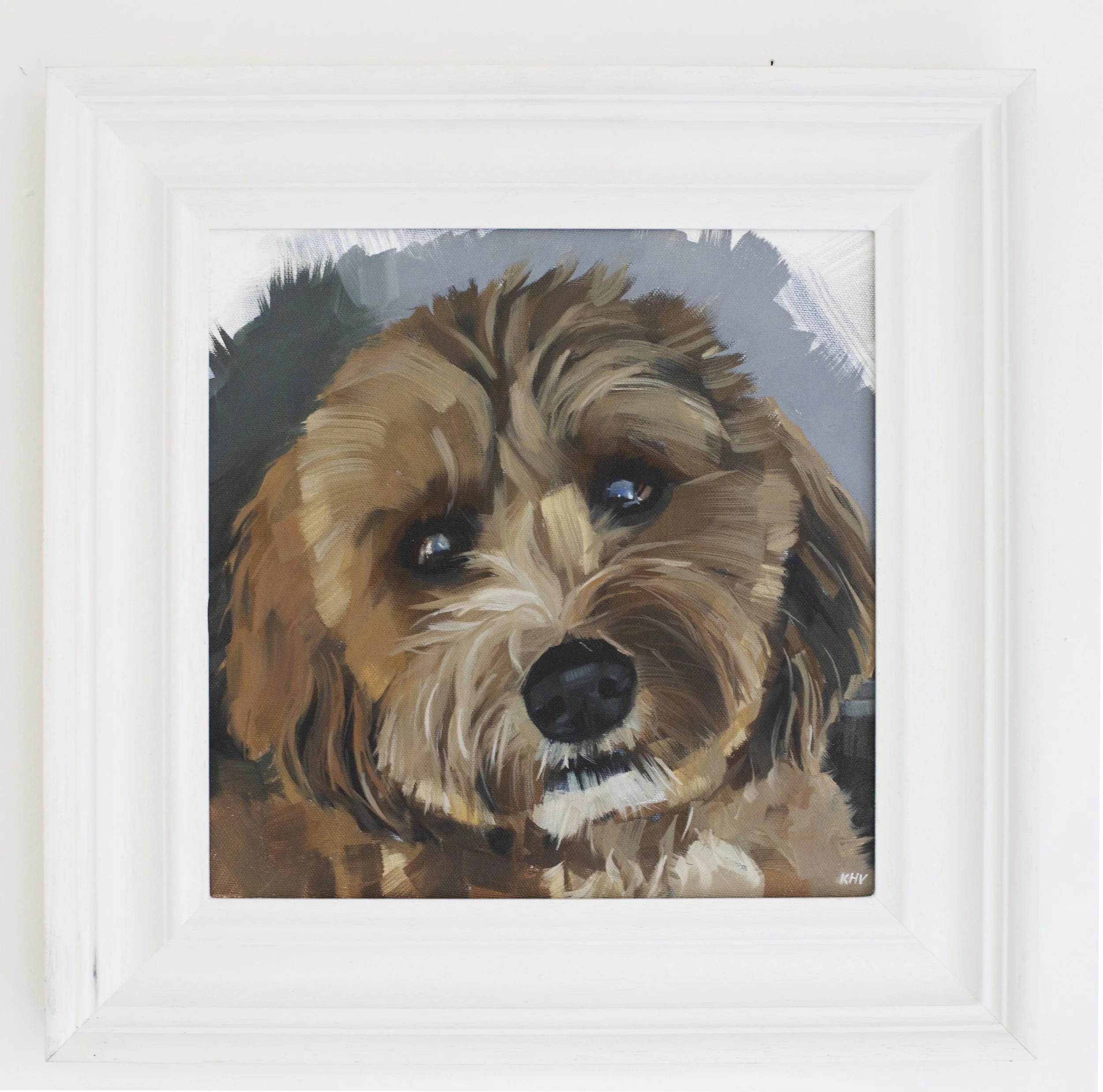 Close-up of an framed expressive oil painting of a cockapoo dog with brown fur, and shiny eyes, signed with initials 'KHV' in the bottom corner.