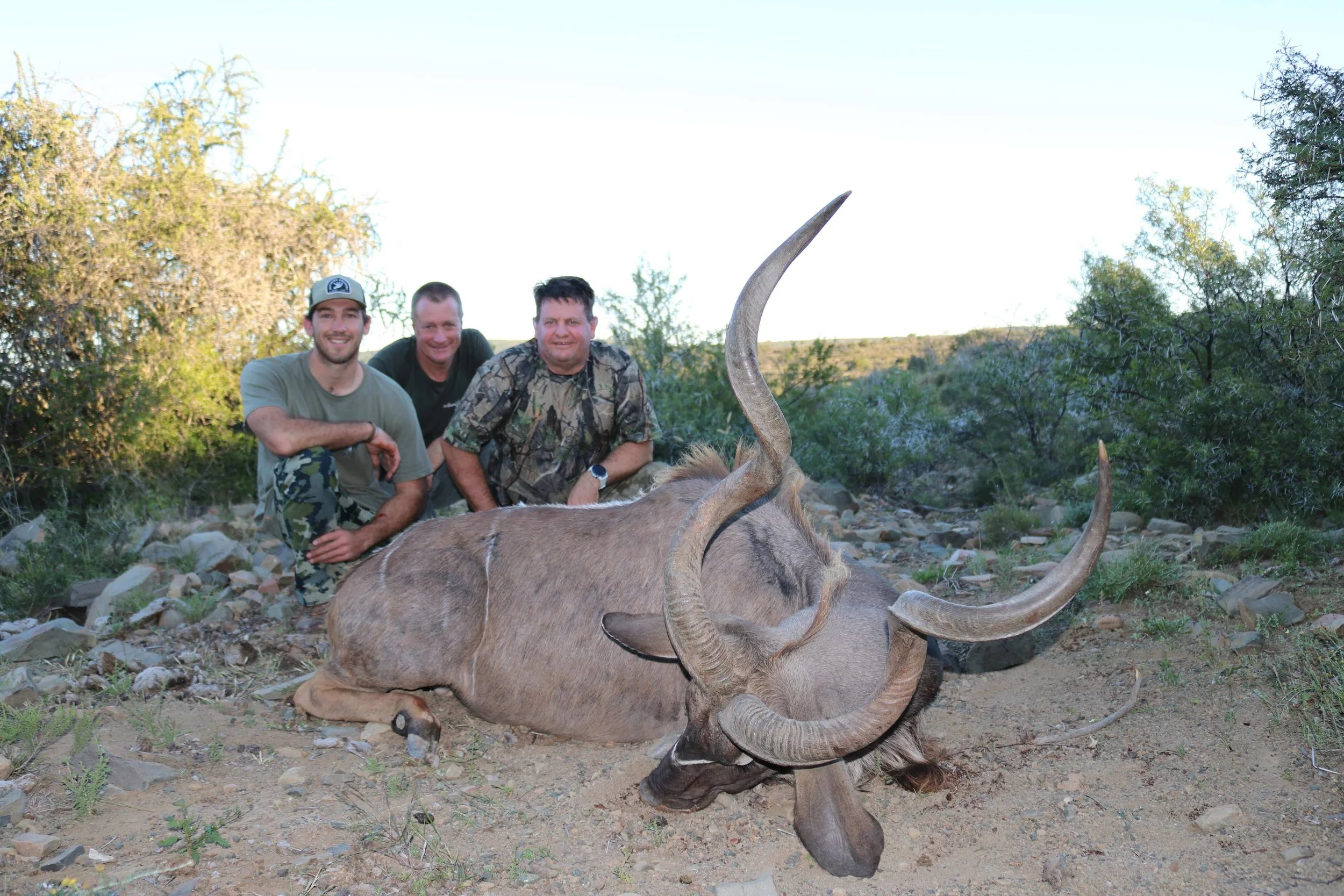 Three men posing behind a dead large antelope with twisted horns, lying on the ground in a desert-like area with bushes and rocky terrain.