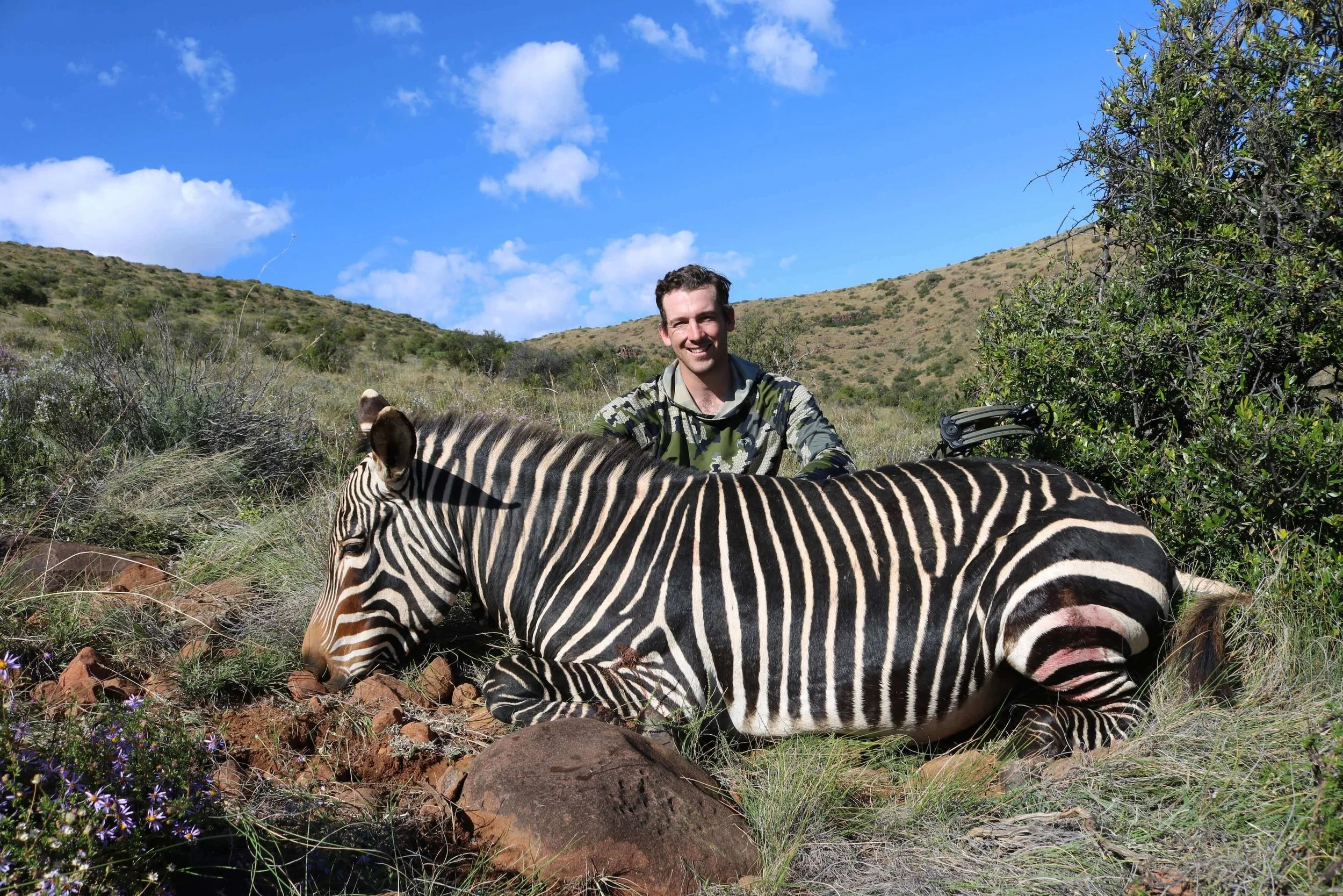 A man smiling next to a sleeping zebra in a grassy, hilly landscape under a blue sky with white clouds.