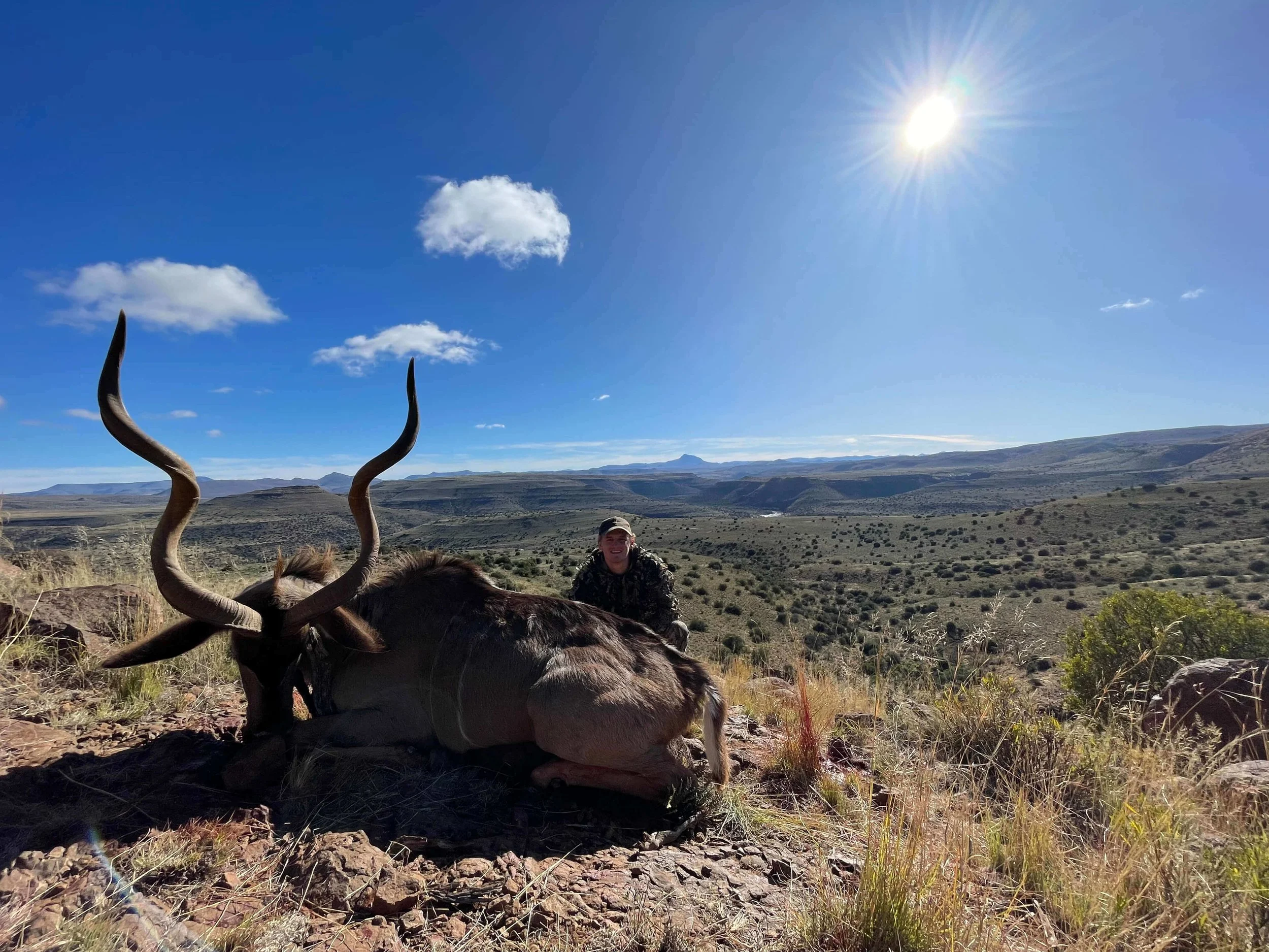 A person kneeling next to a large dead antelope with long, curved horns in a vast, open desert landscape under a bright sun with few clouds.