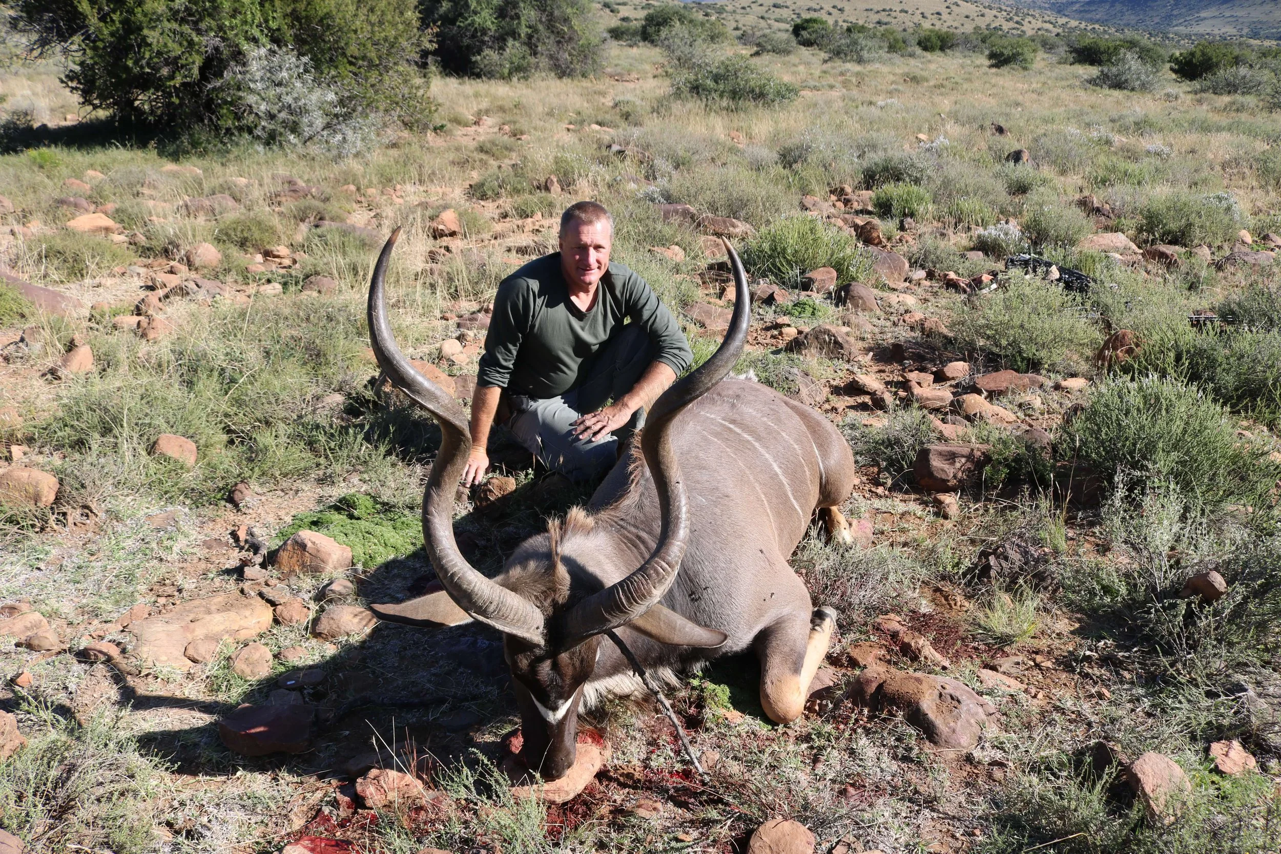 A man kneeling next to a dead kudu with large, curved horns in a desert landscape with scattered rocks and bushes.