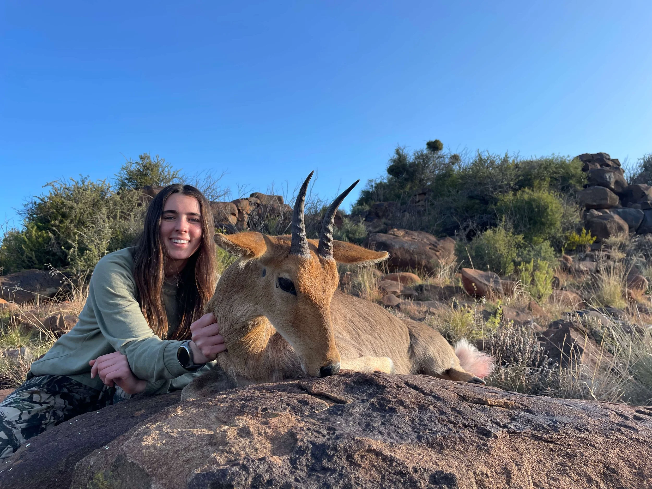 A young woman with long brown hair crouches next to a resting goat with curved horns on a rock in a desert-like landscape with rocks and sparse bushes, under a clear blue sky.