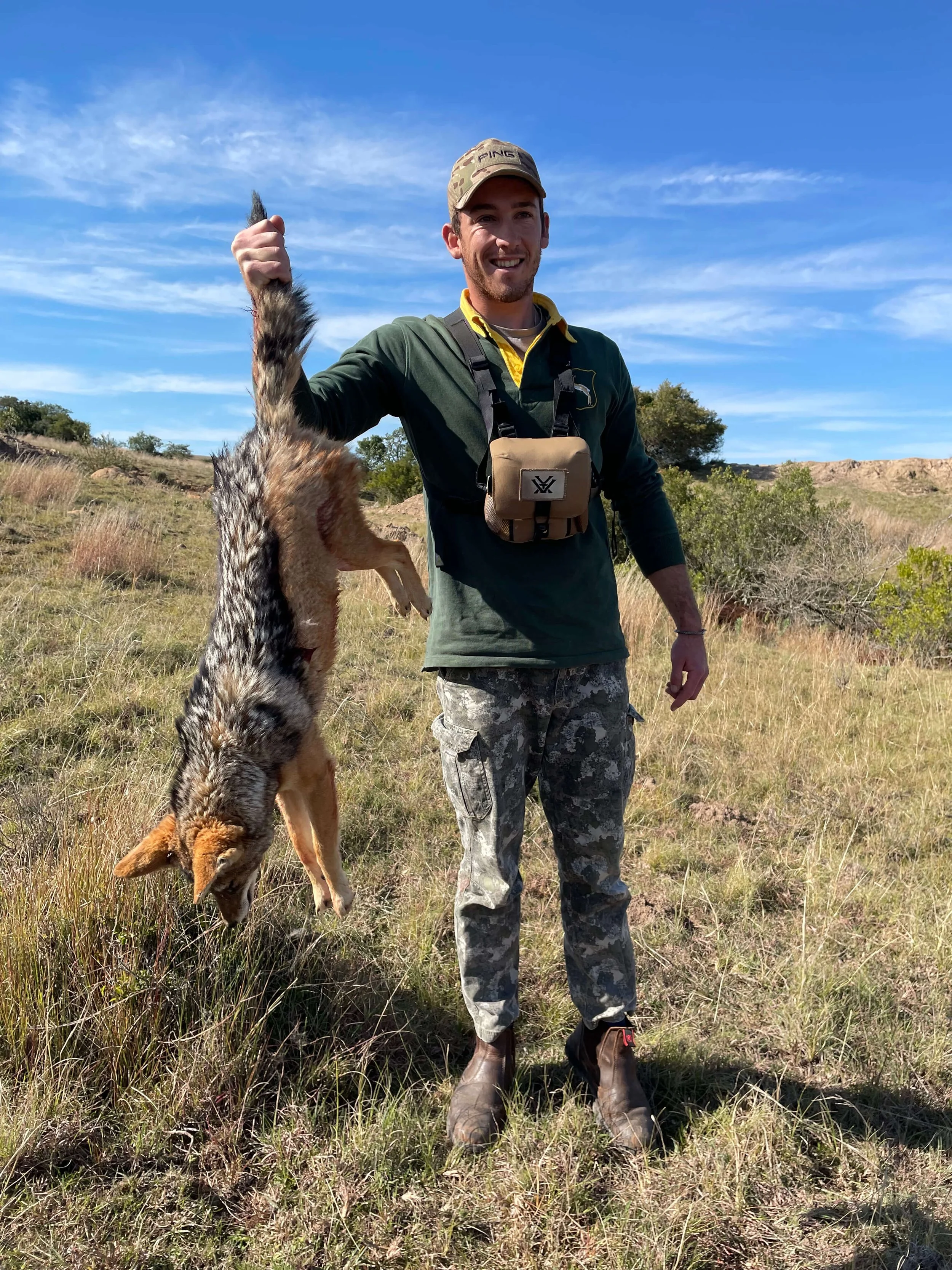 A man in outdoor camo pants, green jacket, and a baseball cap is standing in a grassy field, holding a recently hunted coyote by the tail, smiling at the camera with a bright blue sky in the background.