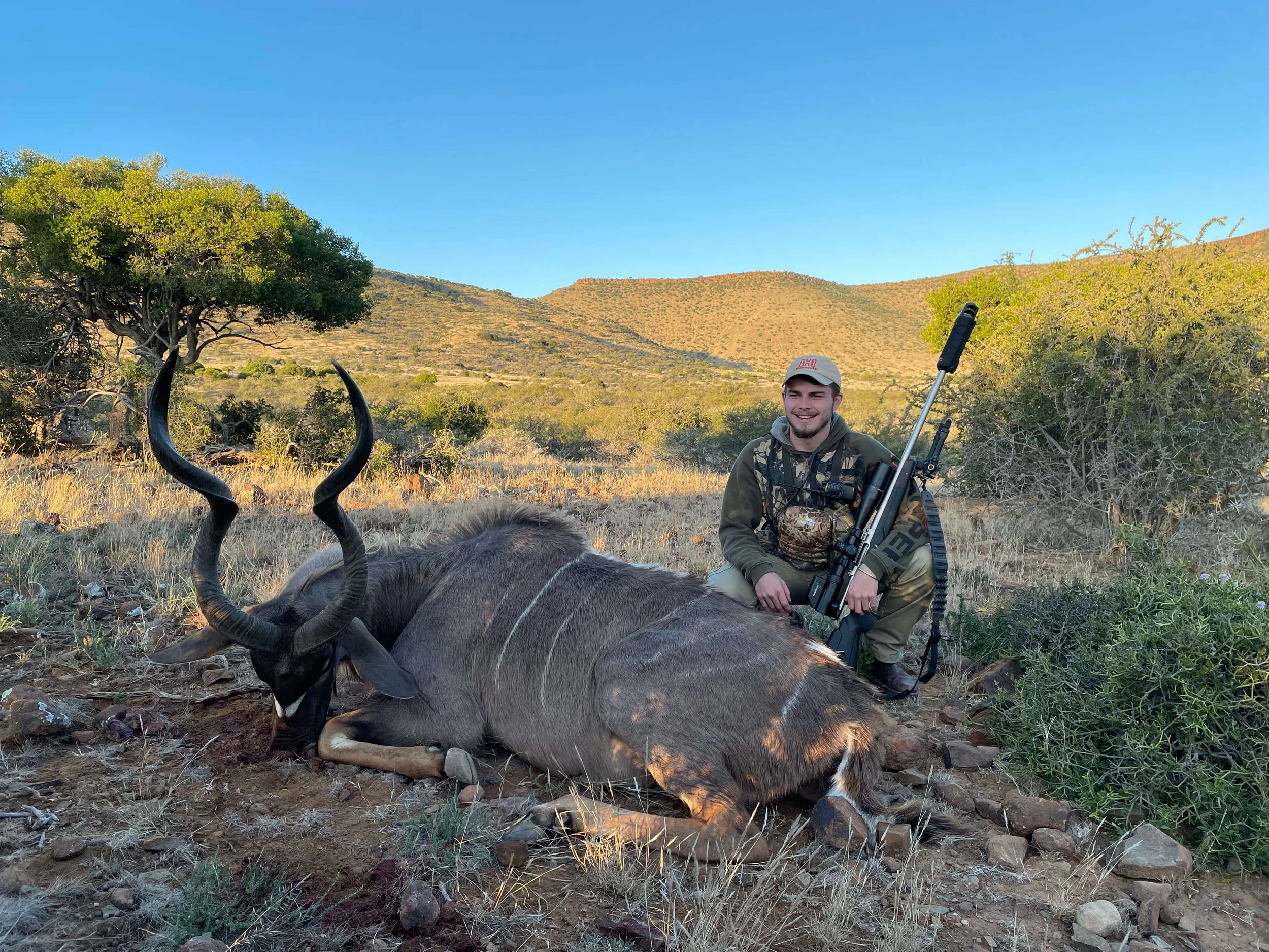 A man kneeling next to a dead antelope with large curved horns in a grassy, hilly landscape with scattered bushes and trees, holding a large rifle.