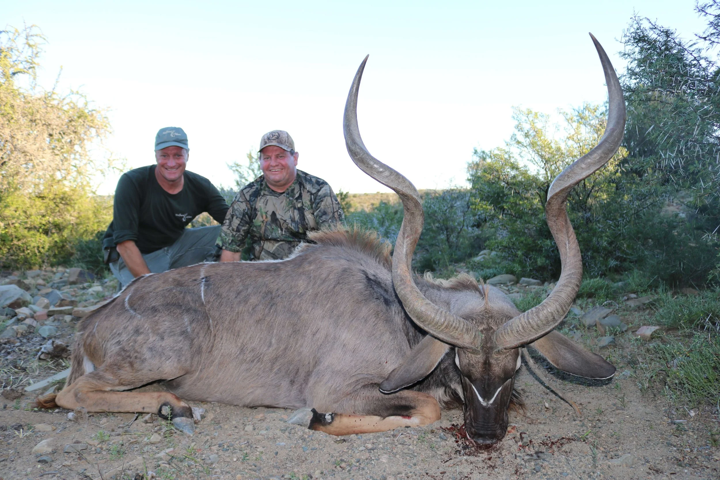Two smiling hunters kneel behind a recently killed large antelope with long, curved horns in a dry, rocky landscape with green bushes and trees.