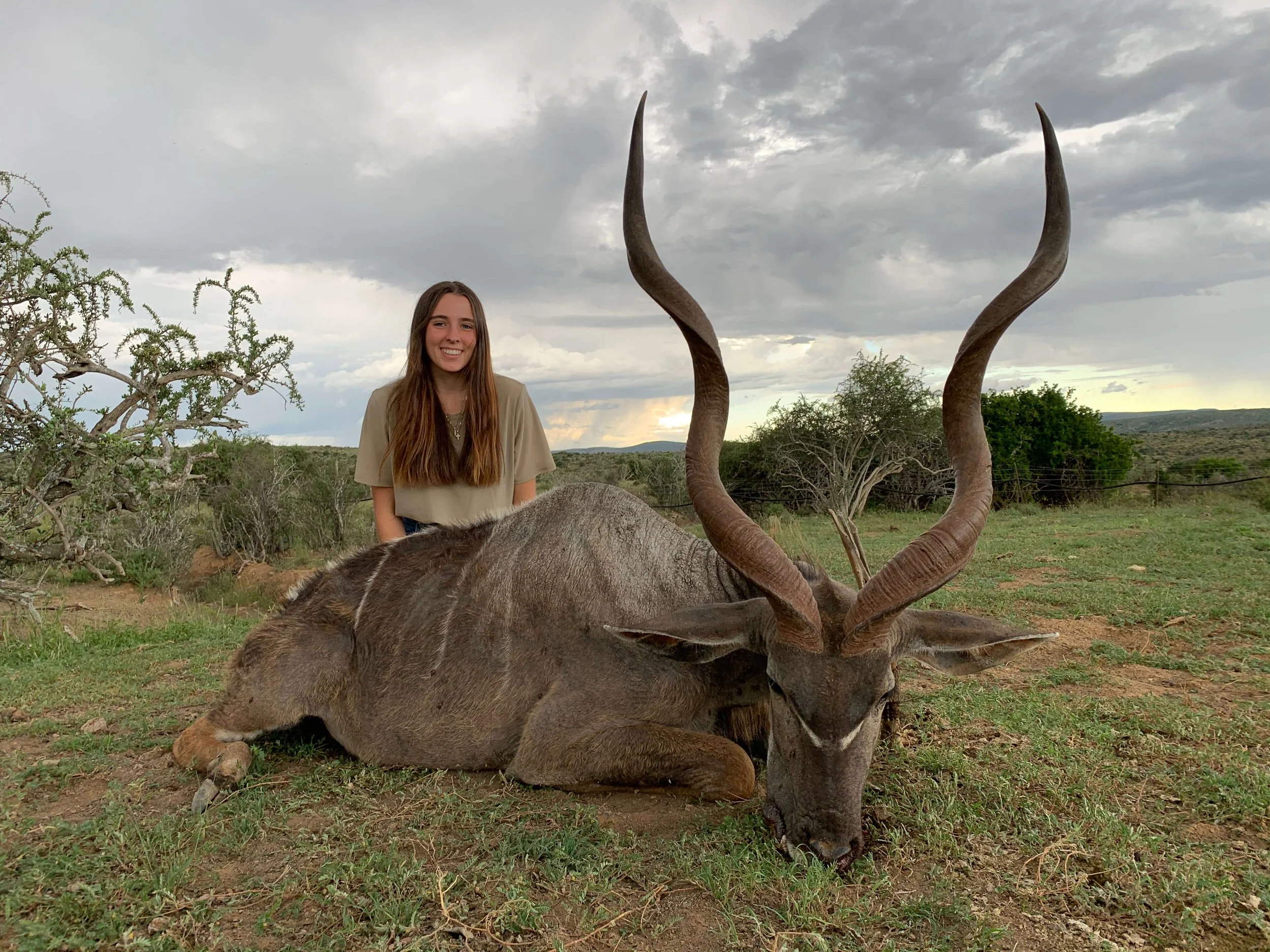 A young woman with long brown hair smiling next to a dead kudu antelope lying on the ground, with a cloudy sky and sparse trees in the background.