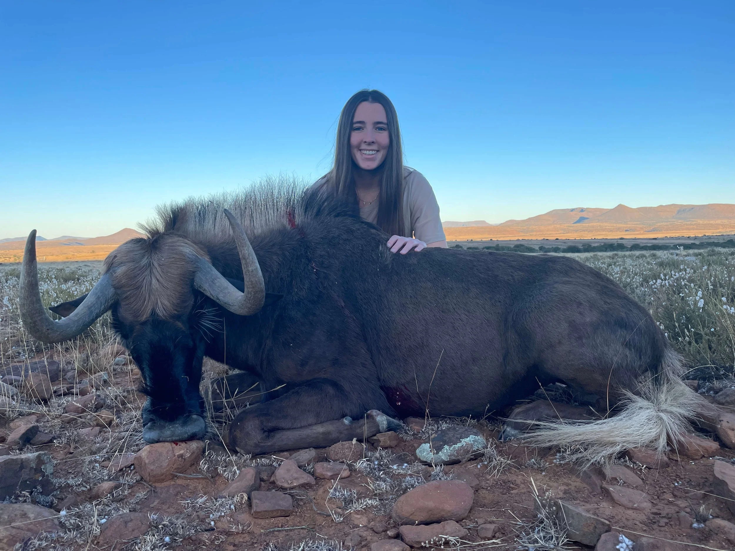 A woman with long hair smiling and resting her hand on a large dead wildebeest lying on rocky ground in an open grassland with mountains in the background under a clear blue sky.