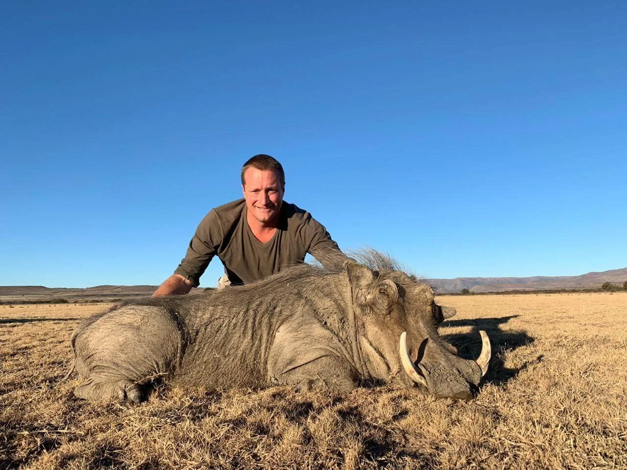 A man in a brown shirt kneeling next to a large dead elephant lying on dry grassland.