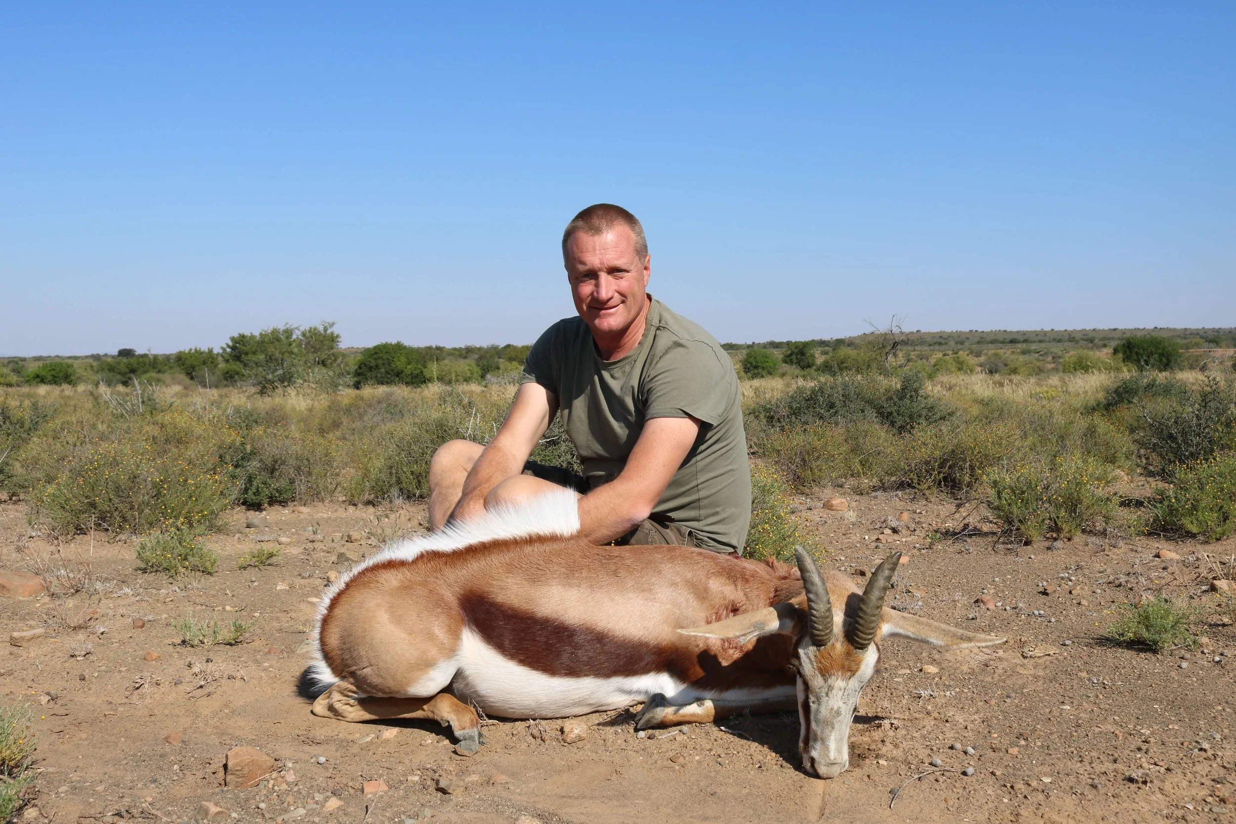 A man kneeling in a desert landscape next to a dead gazelle with curved horns.