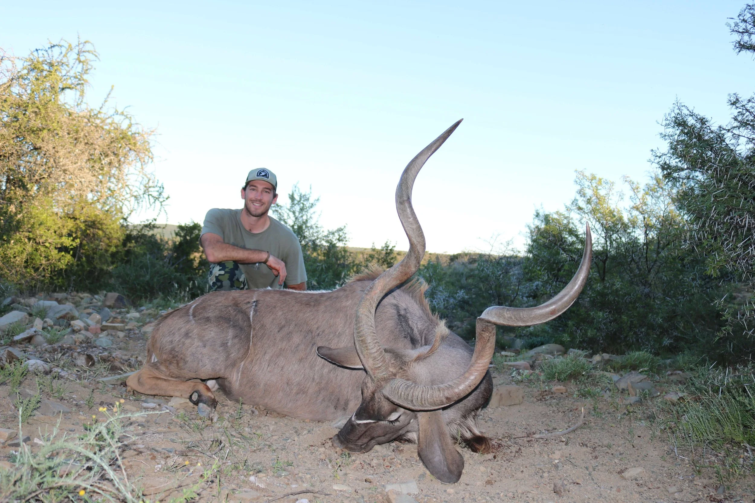 A man kneeling next to a large, dead antelope with long, curved horns in a natural outdoor setting with bushes and trees.
