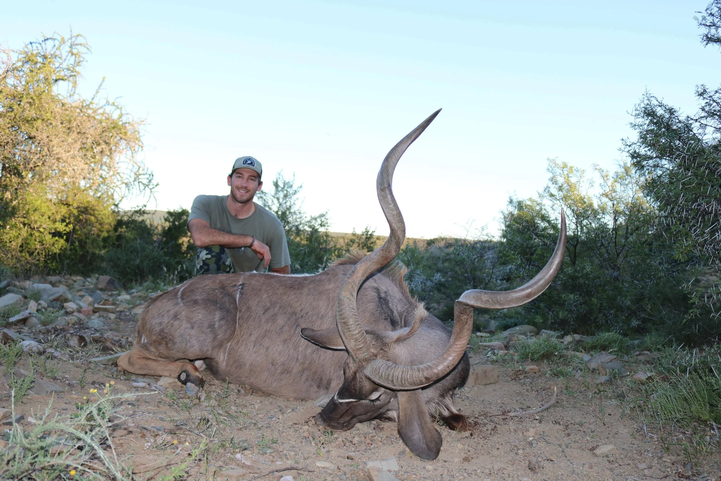 A man crouching beside a recently hunted large antelope with long, curved horns in a natural, bushy landscape under a clear sky.