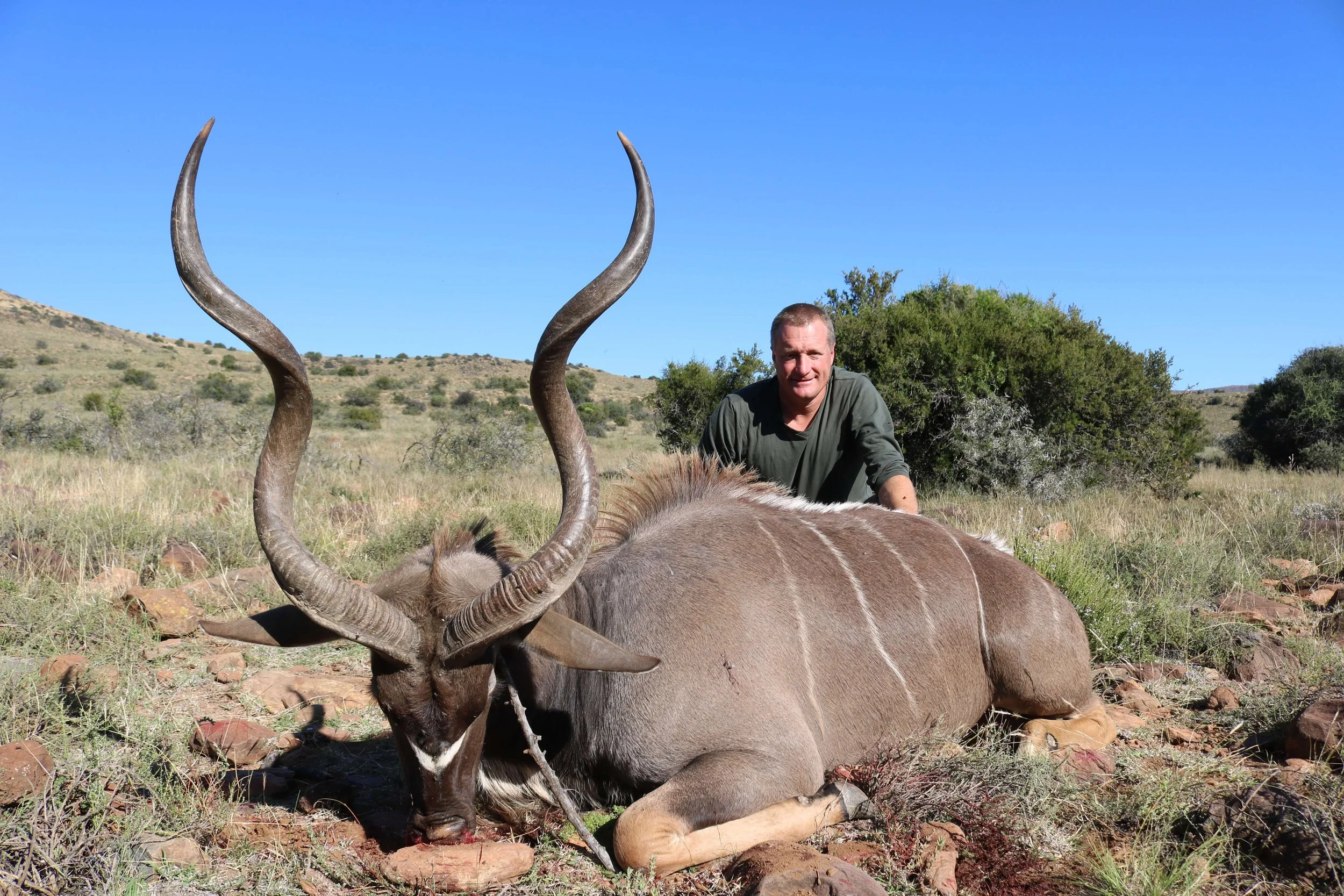 A man kneeling behind a dead kudu with curved horns in a dry, grassy landscape with bushes and hills in the background.
