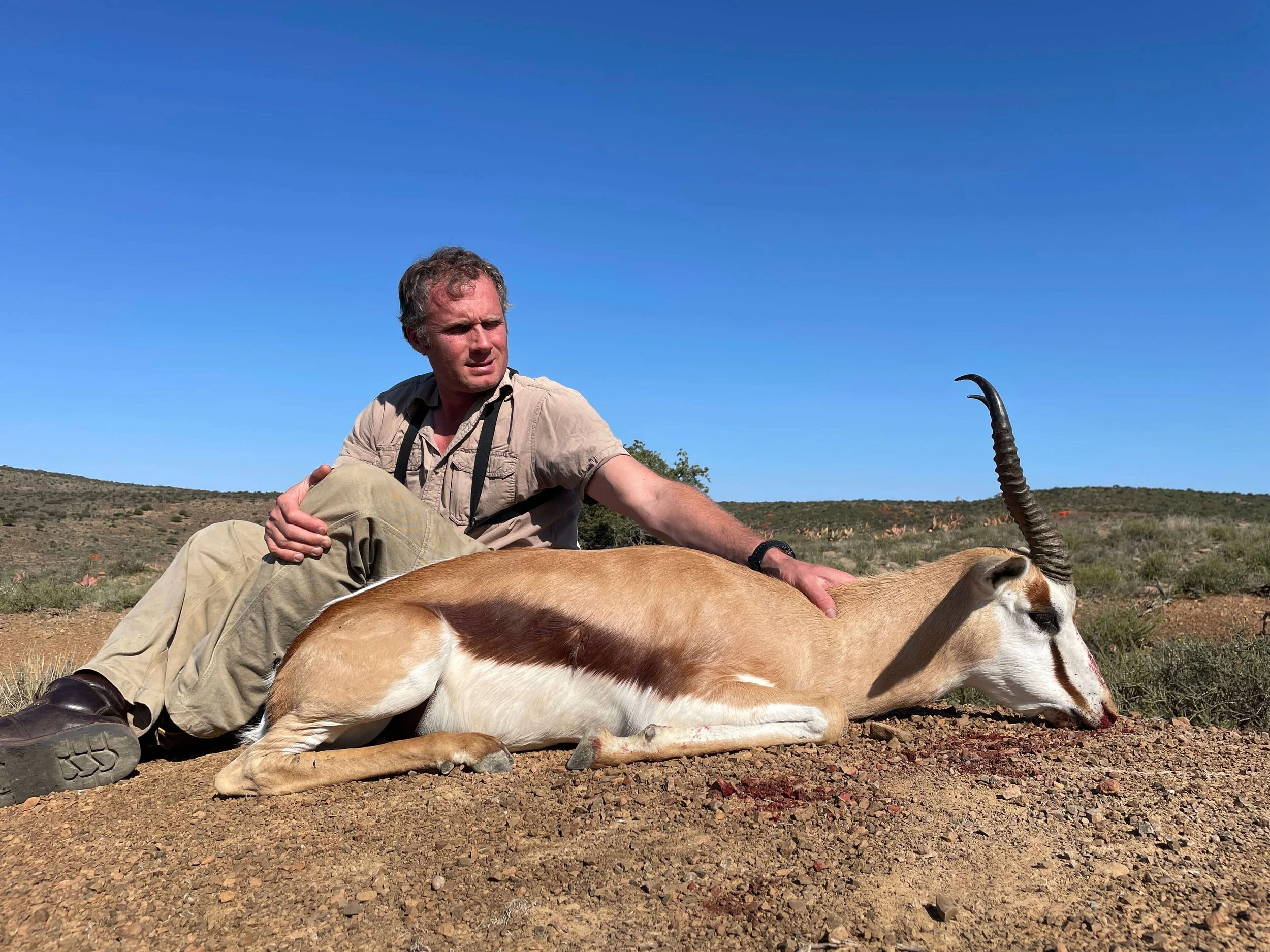 A man in beige attire with a camera around his neck sitting on desert ground next to an oryx with long, twisted horns, lying down with its head resting on the ground, in a semi-arid landscape under a clear blue sky.