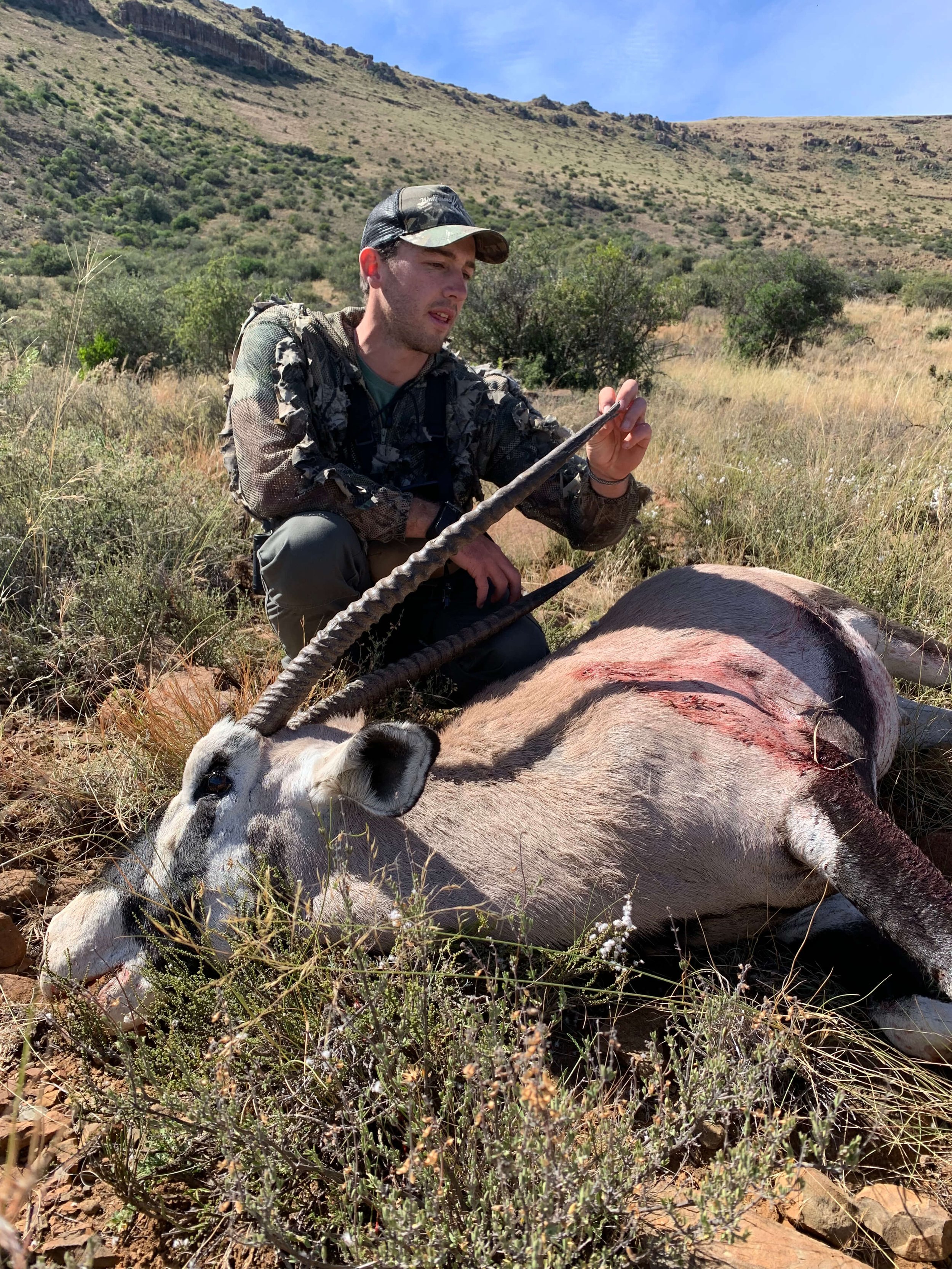 Young man wearing camouflage outdoors, kneeling next to a dead oryx with a wound, in a grassy, hilly landscape.