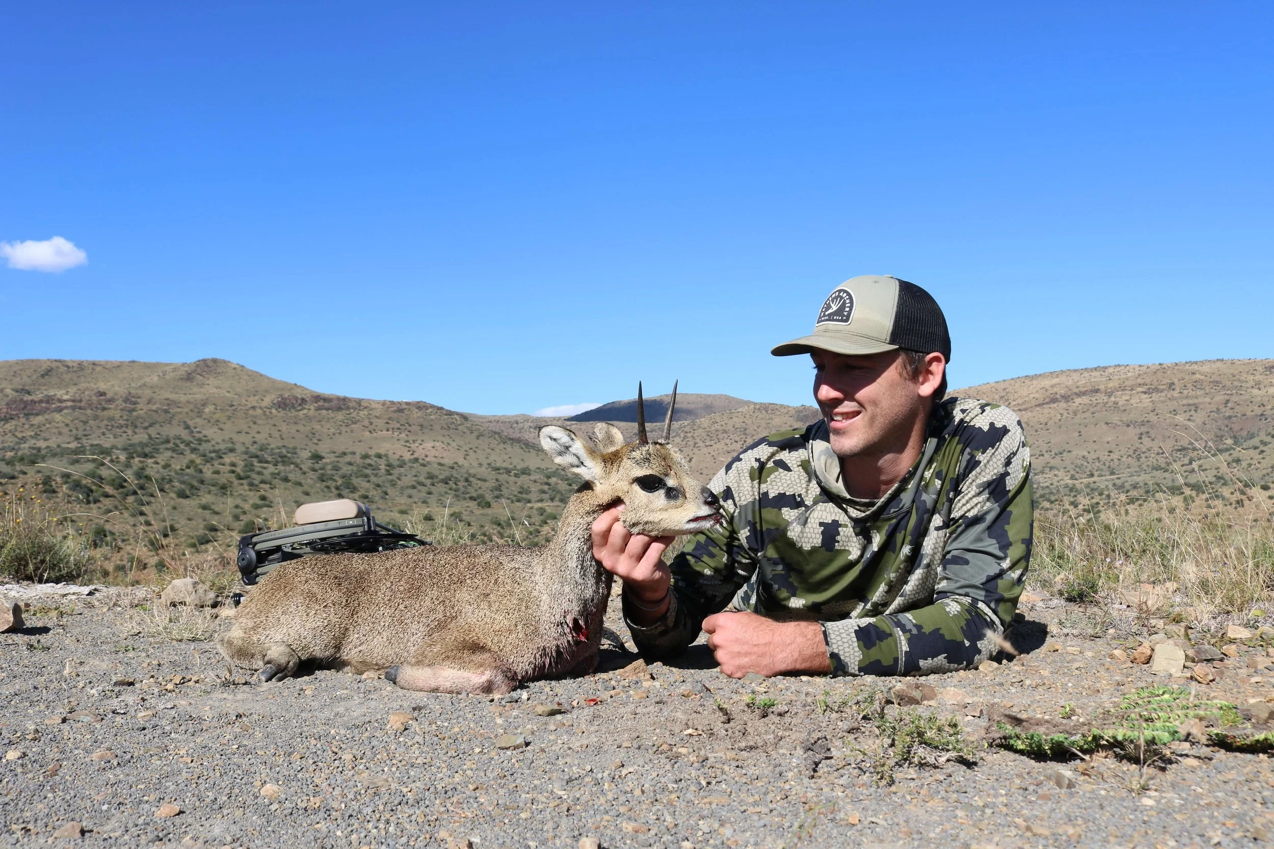 A man lying on the ground in a desert landscape holding a small antelope's head with one hand, with a bow and colored quiver next to him, under a blue sky with some clouds.