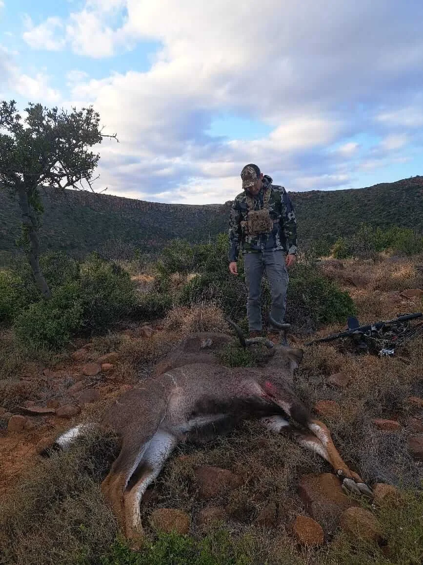 A man in camouflage and outdoor gear standing next to a deceased pronghorn antelope in a desert landscape with hills, shrubs, and a partly cloudy sky.