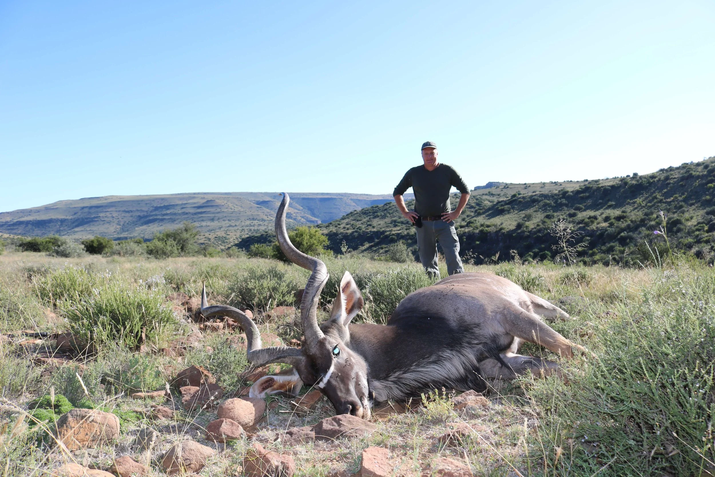 A man standing in a grassy field with his hands on his hips, behind a dead antelope with long, curved horns, lying on the ground. The background shows hills and clear blue sky.