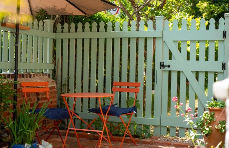 A cozy brick patio with a small, round orange table and two matching chairs, shaded by an umbrella. Blue and terracotta potted plants decorate the space. The area is enclosed by a light blue wooden picket fence and gate.
