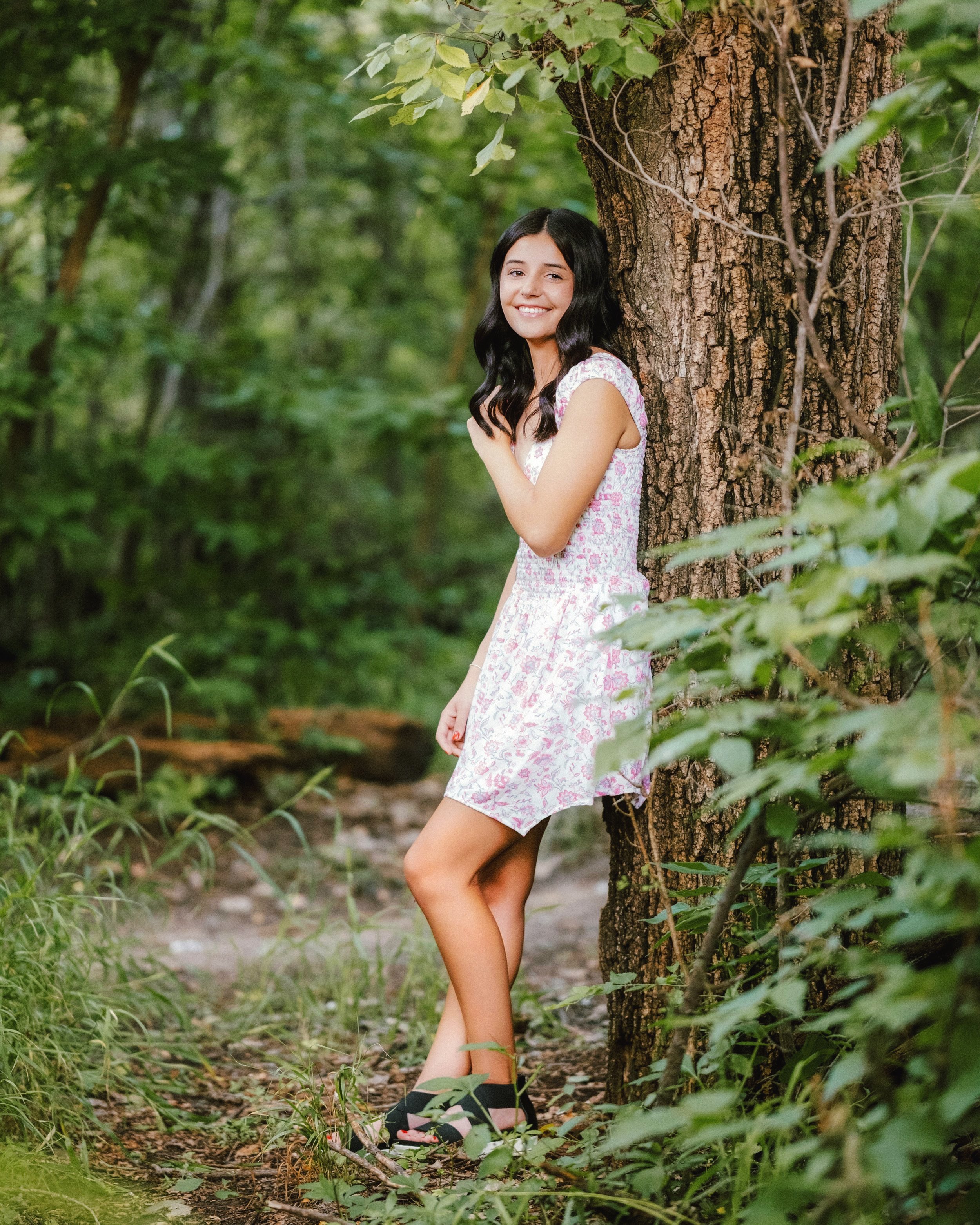 A young woman with black hair wearing a white floral dress and black sandals leaning against a tree in a green forest.