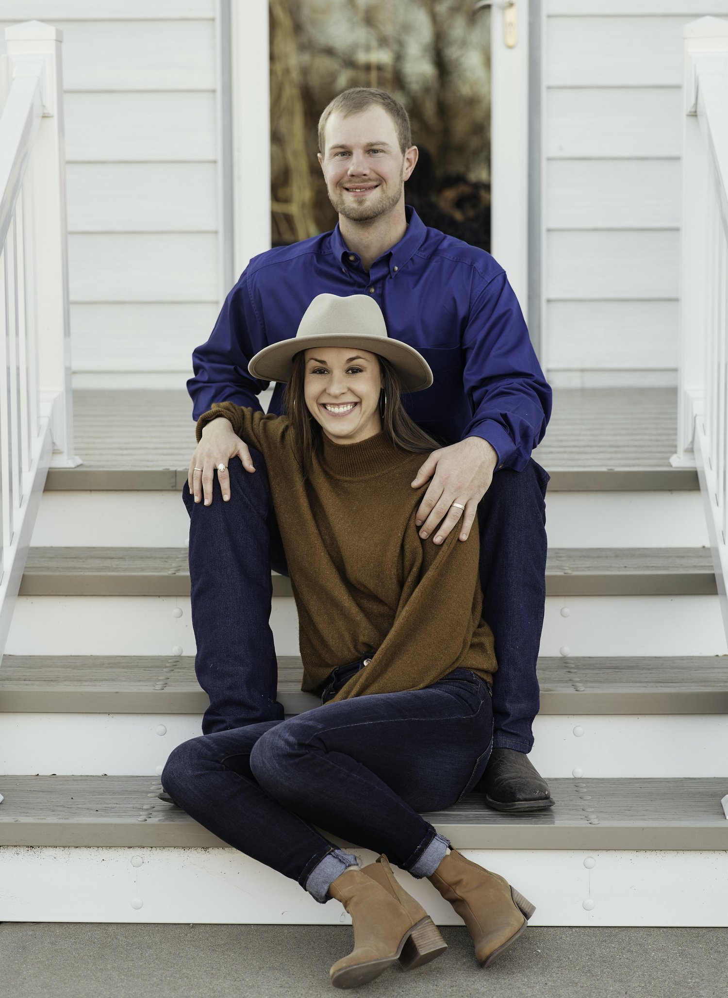 A man and woman sitting on outdoor stairs, smiling, with the man standing behind the woman. The woman wears a wide-brimmed hat, brown sweater, jeans, and ankle boots. The man wears a blue button-up shirt and dark jeans. The background shows a porch a