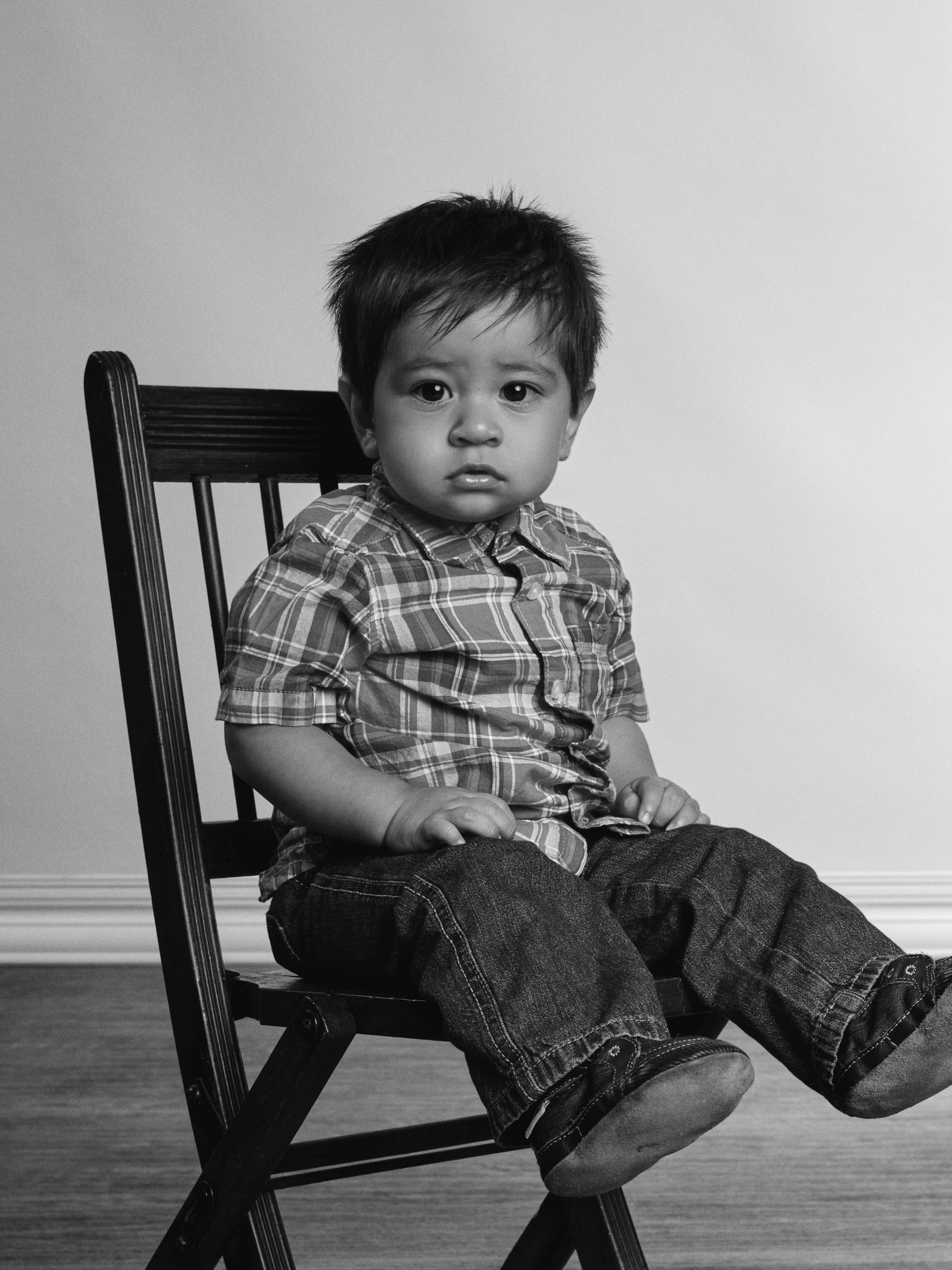 Black and white photo of a young boy sitting on a wooden chair, looking at the camera with a serious expression.