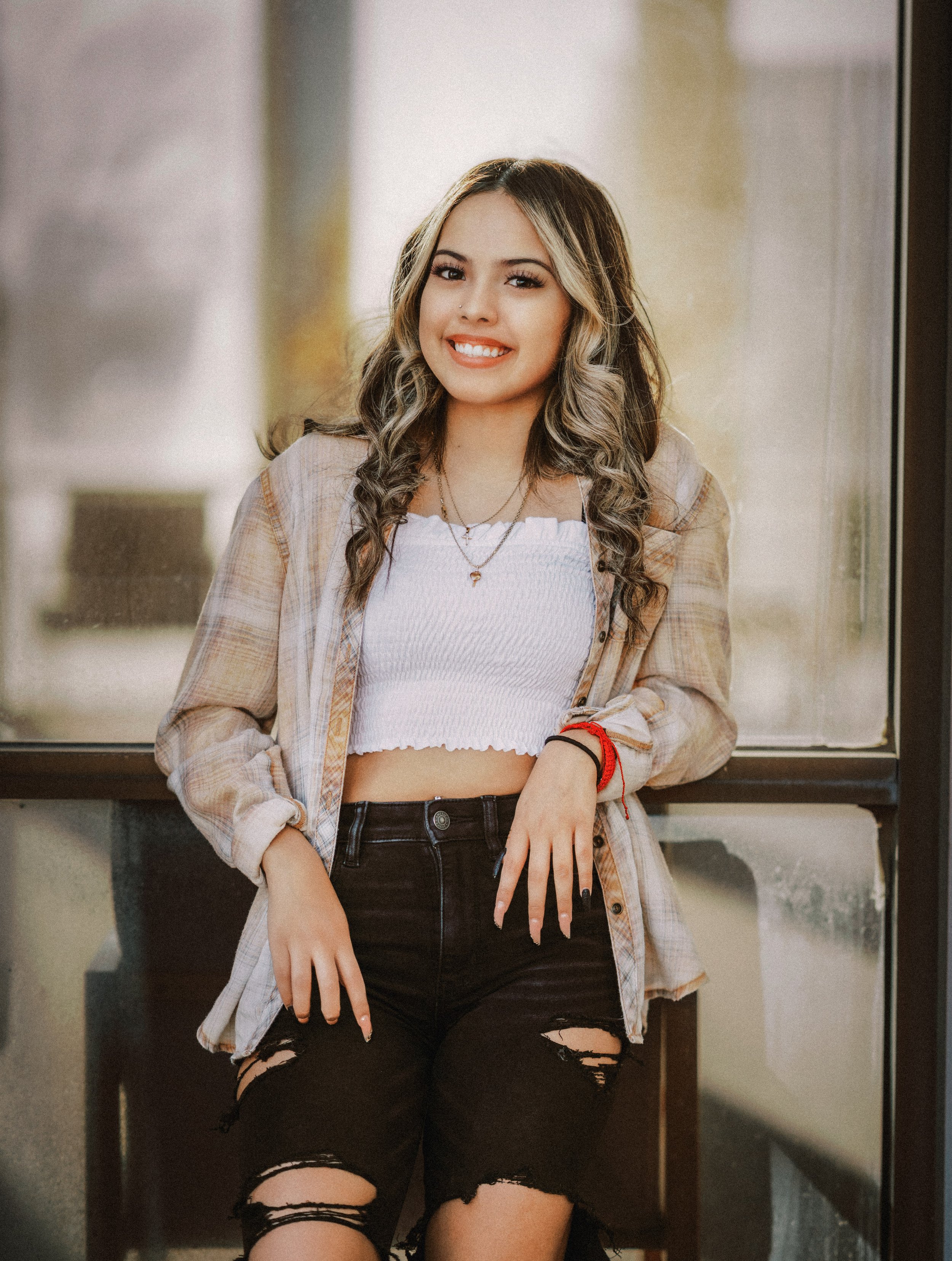 A young woman with wavy hair and a bright smile, wearing a white crop top, a plaid shirt, and ripped black jeans, sitting by a window.
