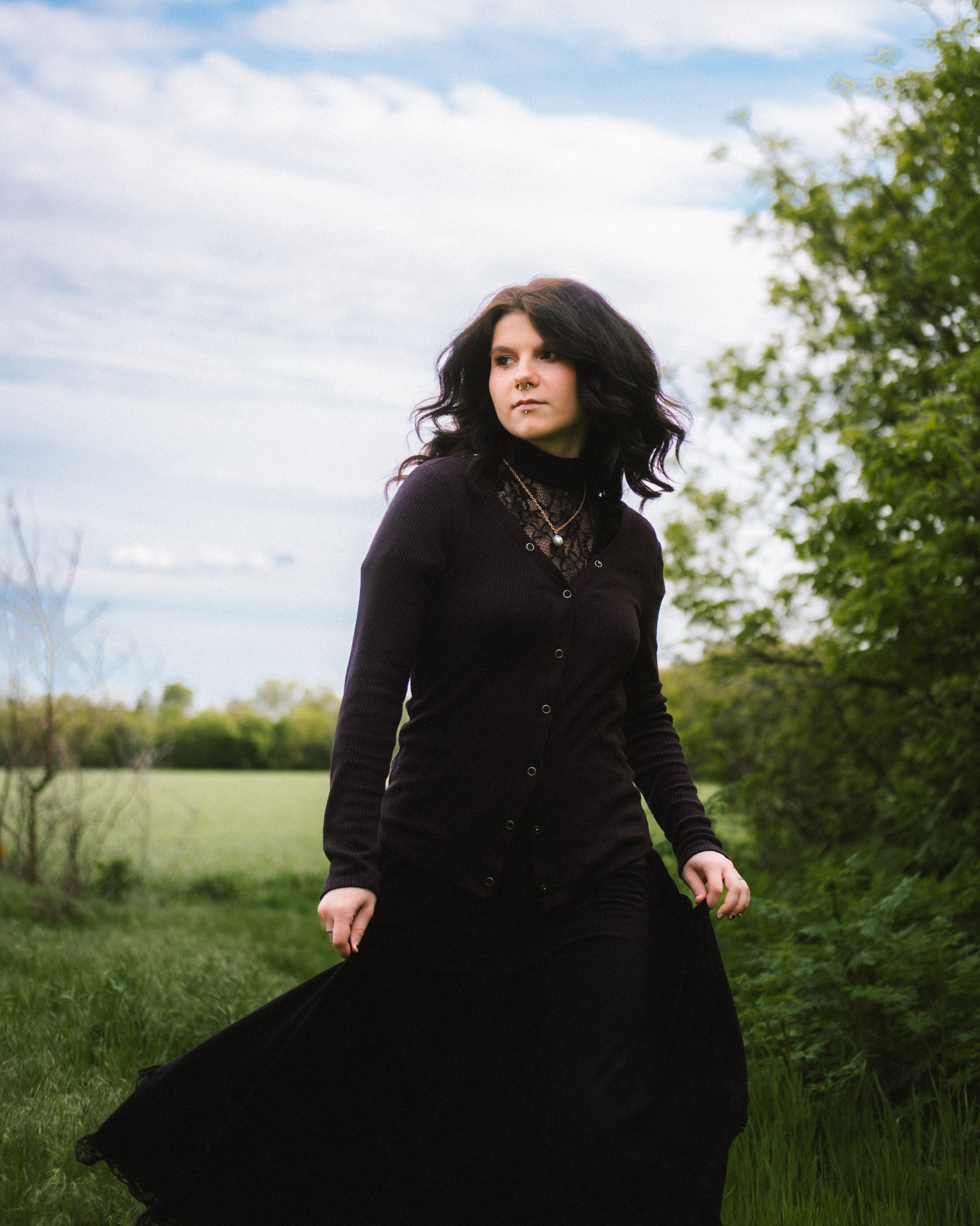 A young woman with dark hair wearing a black cardigan and black skirt, walking outdoors in a grassy area with trees and a cloudy sky in the background.
