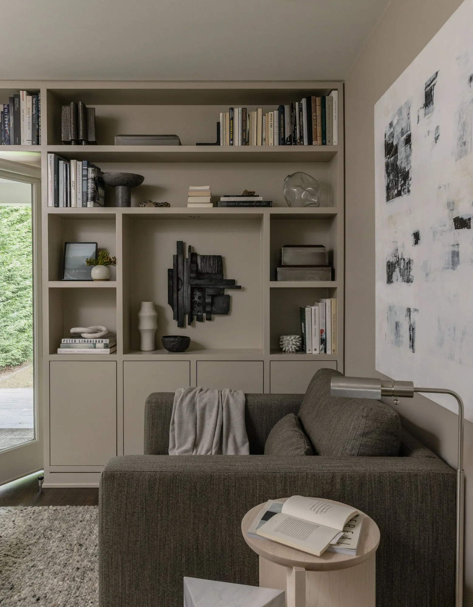 Living room with a gray sofa, a coffee table with an open book, a bookshelf filled with books, decorative objects, and abstract art on the wall.