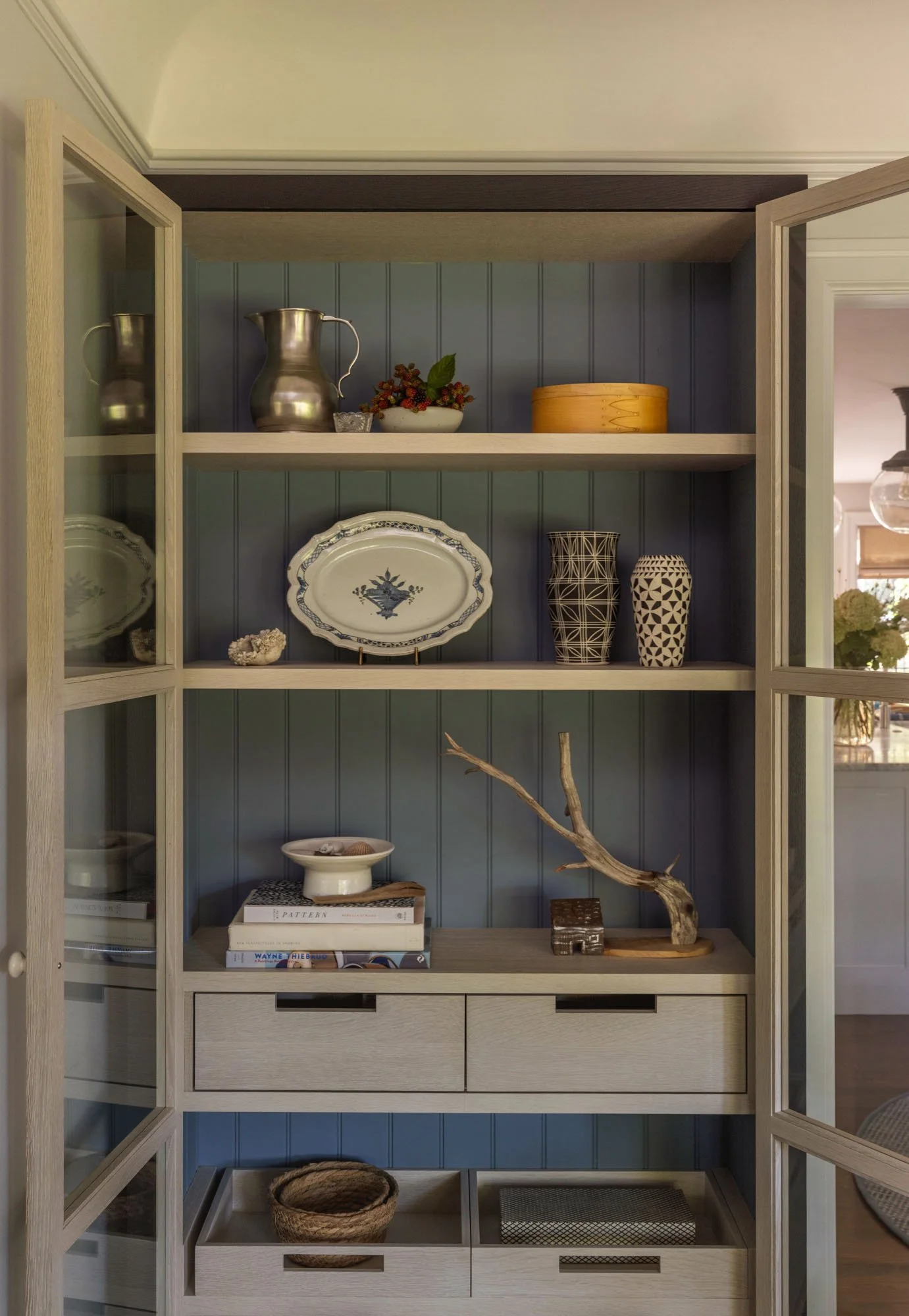 A light wood and glass cabinet with open doors displays decorative plates, ceramic vases, a ceramic bowl, a branch sculpture, books, and woven baskets inside. The background features a blue-green paneled wall.