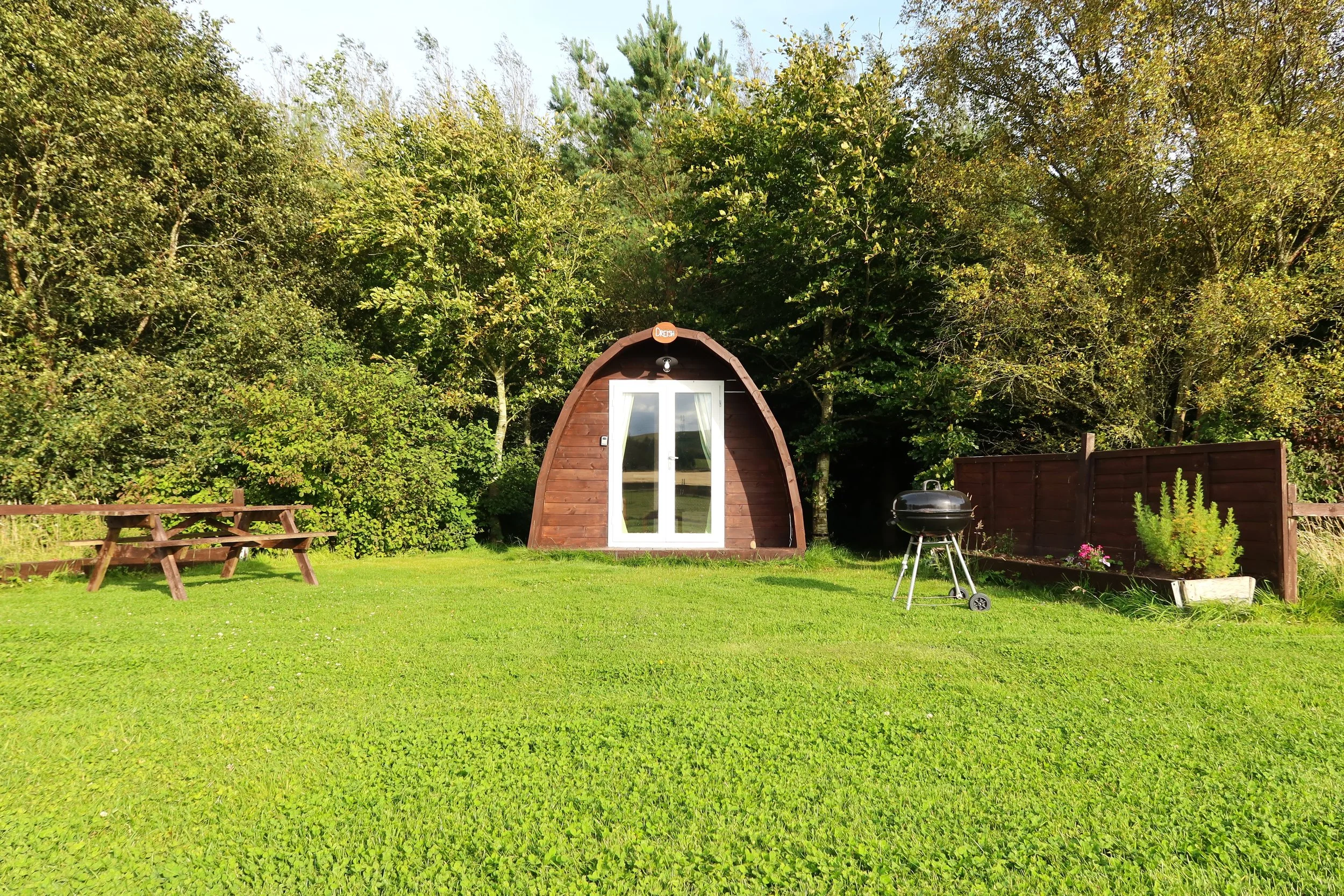 A small wooden igloo-shaped house with a glass door, situated in a green backyard with trees, a picnic table, a barbecue grill, and a wooden fence.