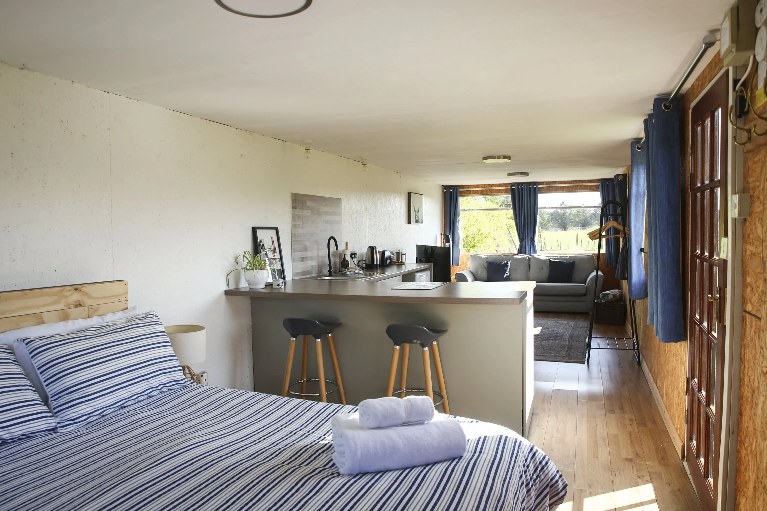 Interior of a cozy studio apartment with a bed, small kitchen, and sitting area with a couch and large window.