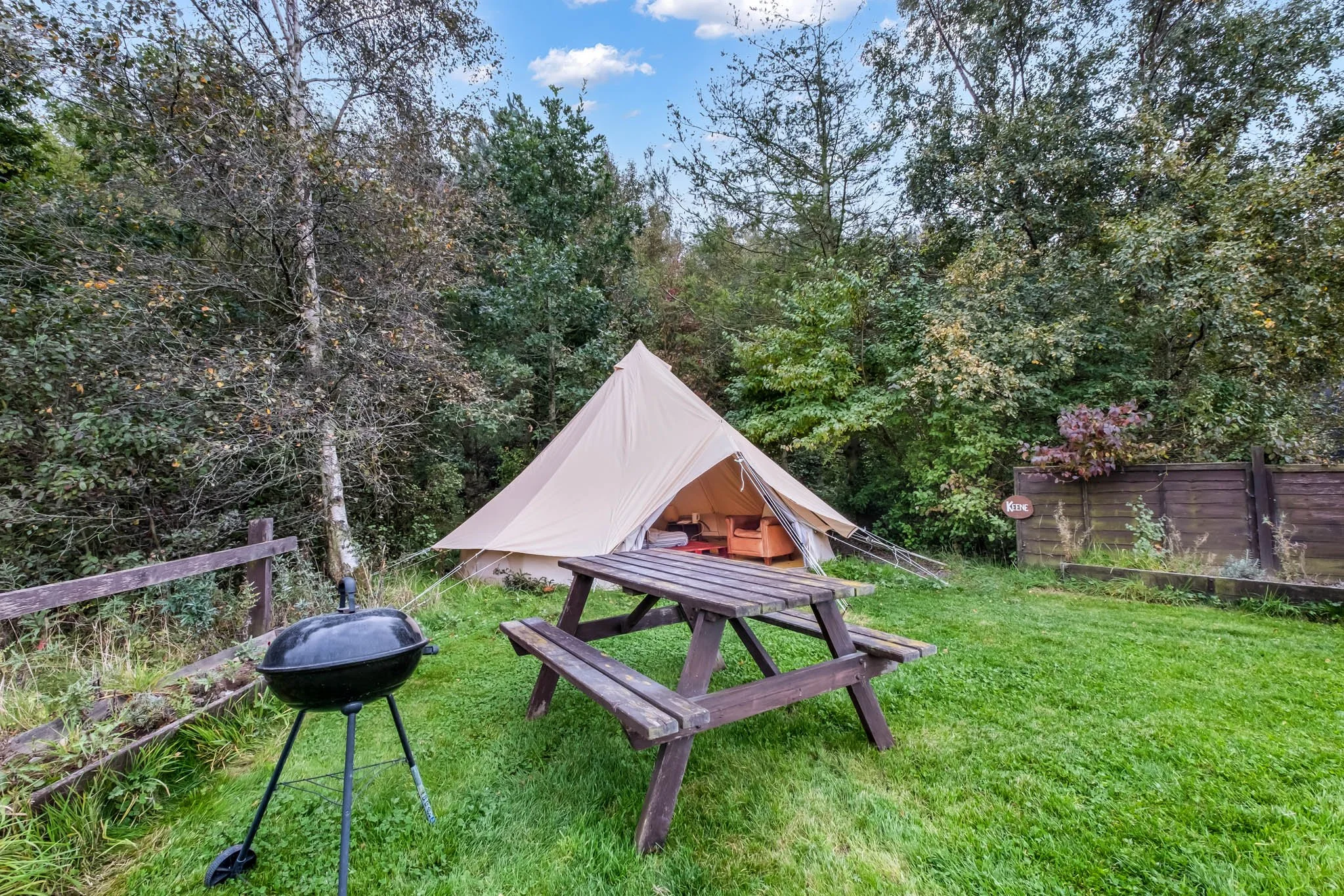 Outdoor backyard scene featuring a beige tent set up among trees, a wooden picnic table, and a black charcoal grill on a grassy lawn with a wooden fence in the background.