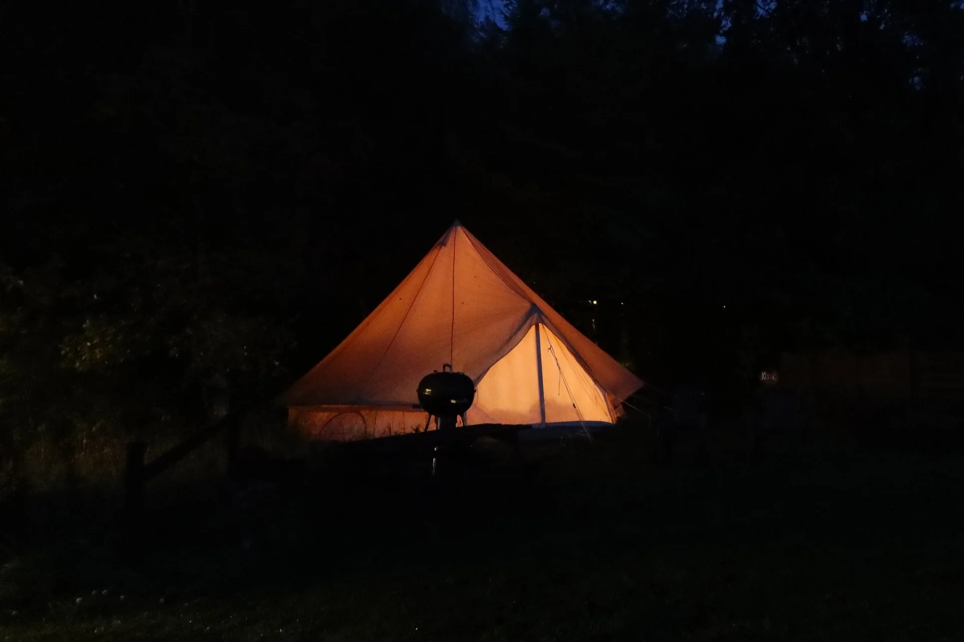 A glowing illuminated tent outdoors at night with a small grill in front of it, surrounded by darkness and trees.