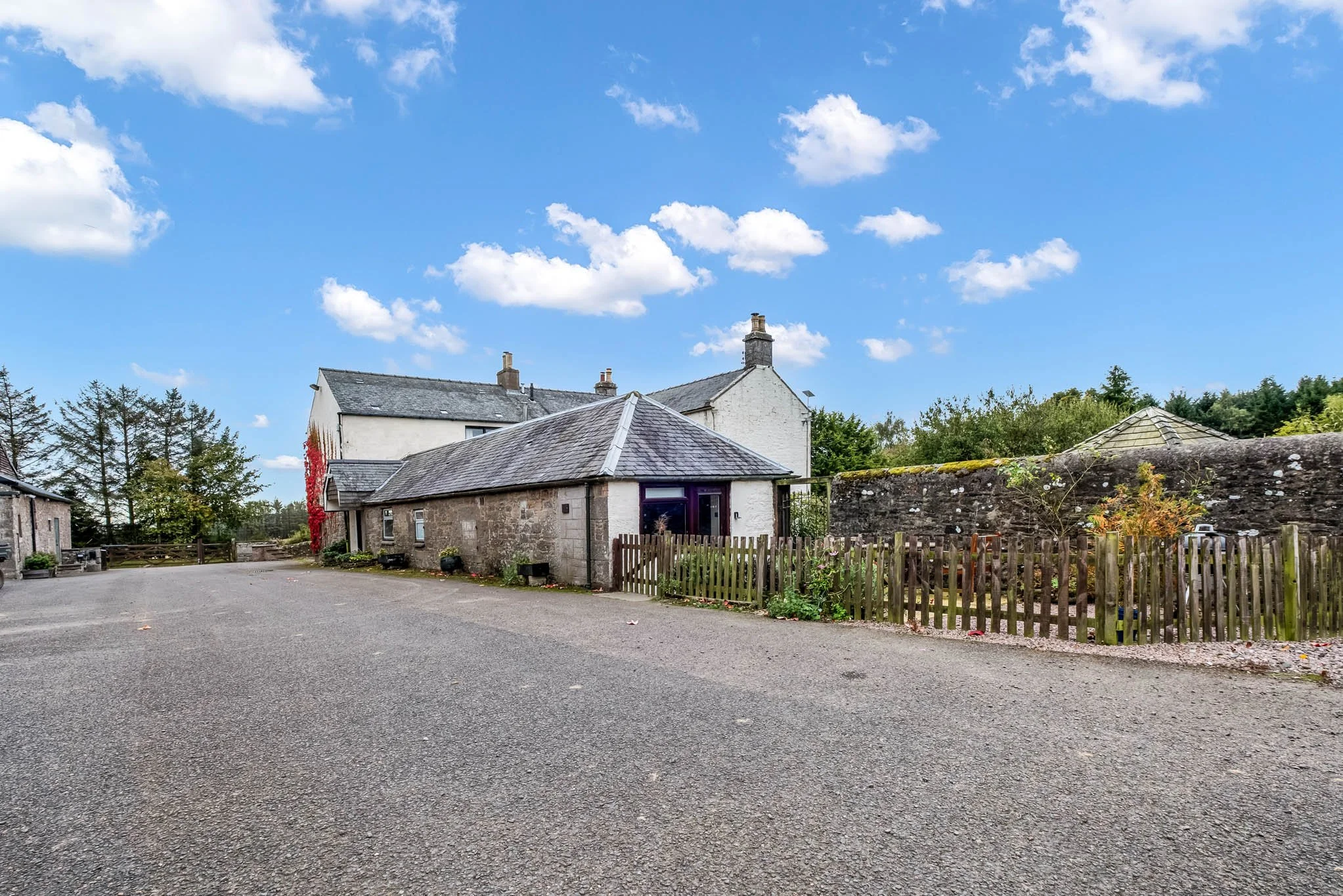 A rural scene with white and stone houses, gravel driveway, wooden fence, and trees under a blue sky with clouds.