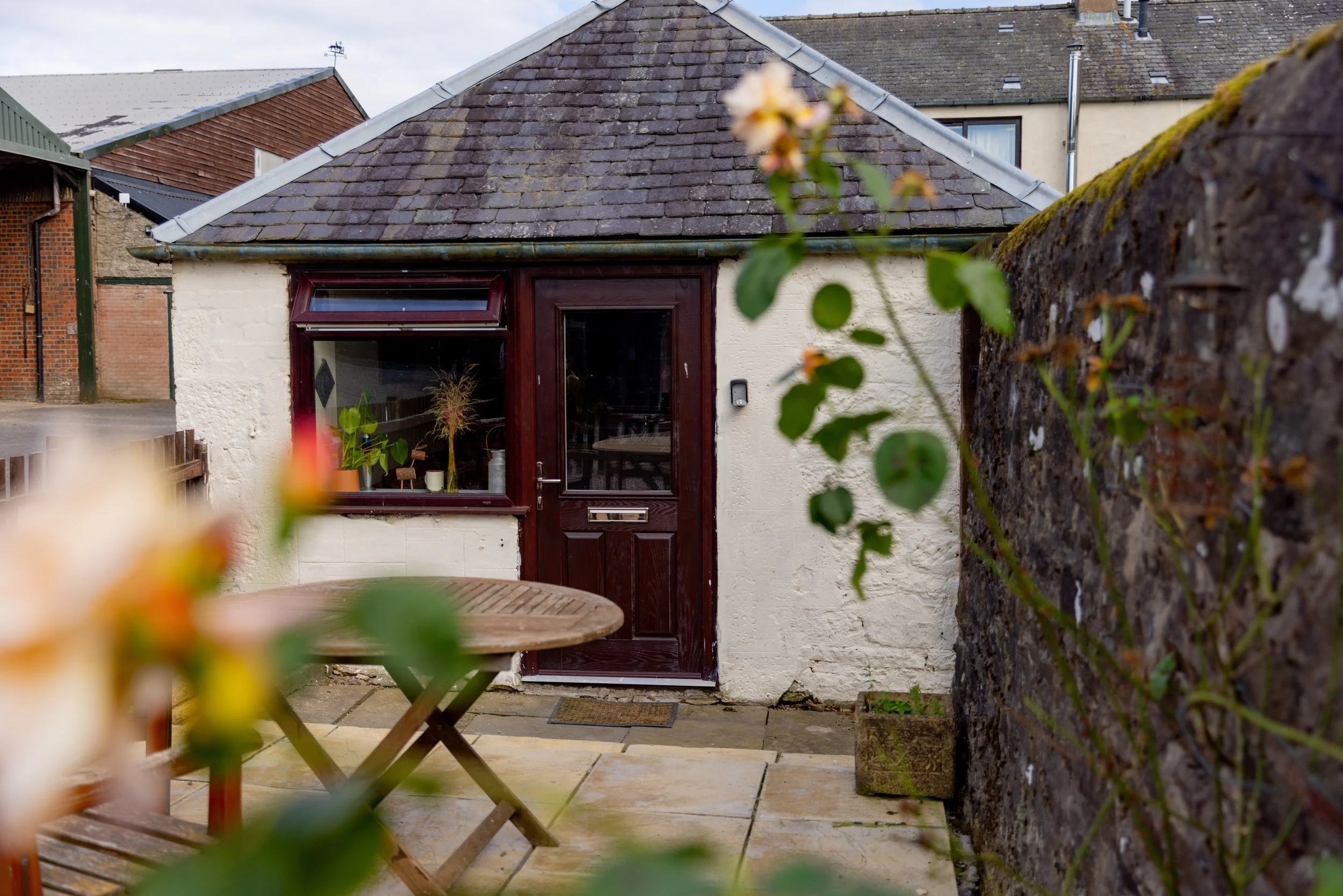 Small white building with a dark brown wooden door and window, surrounded by a stone-paved patio with a round wooden table and blurred flowers in the foreground, enclosed by a stone wall and neighboring buildings with sloped roofs.