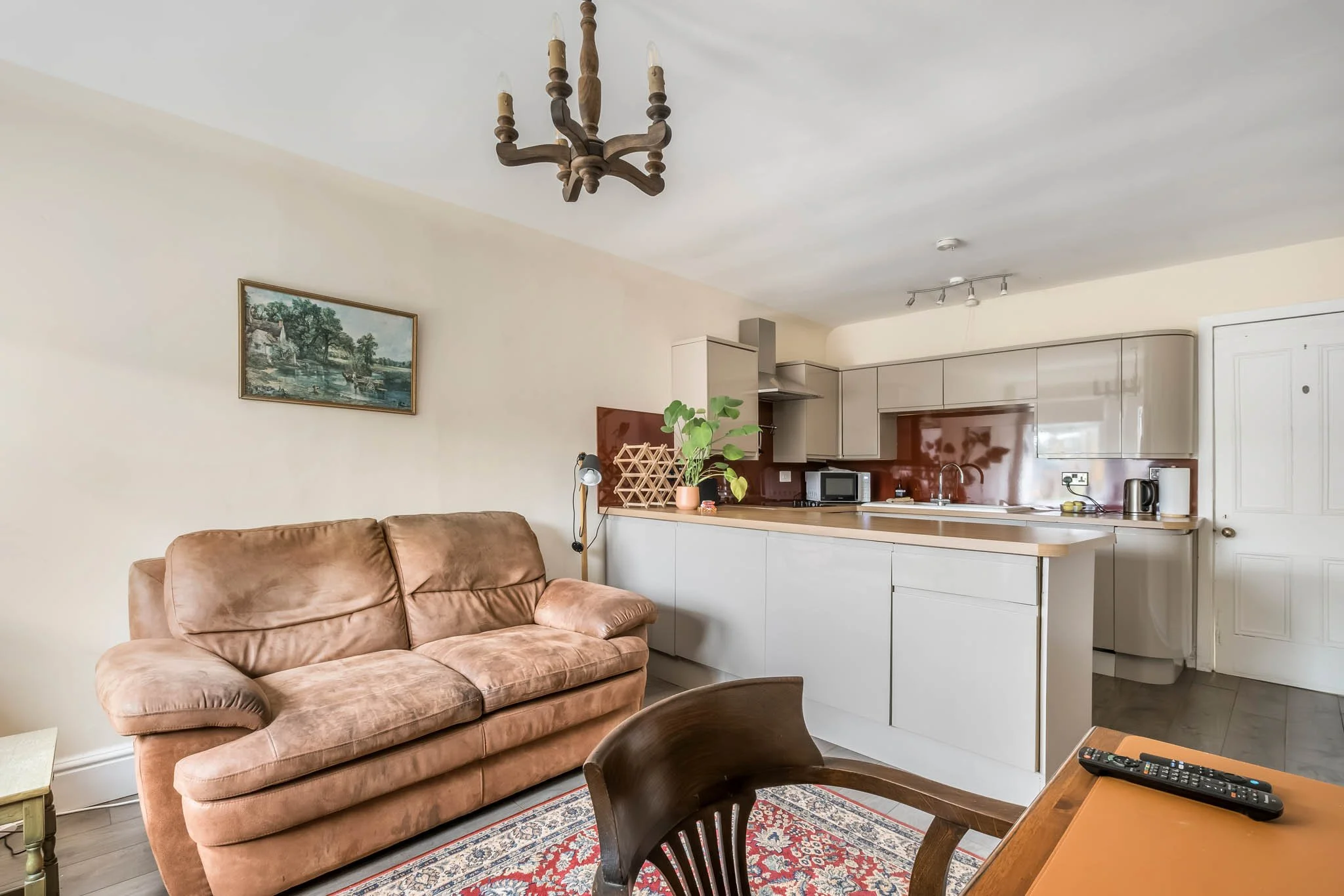Living room with a pink sofa, wooden dining table, and a kitchen with beige cabinets, a small plant, microwave, and a red backsplash.