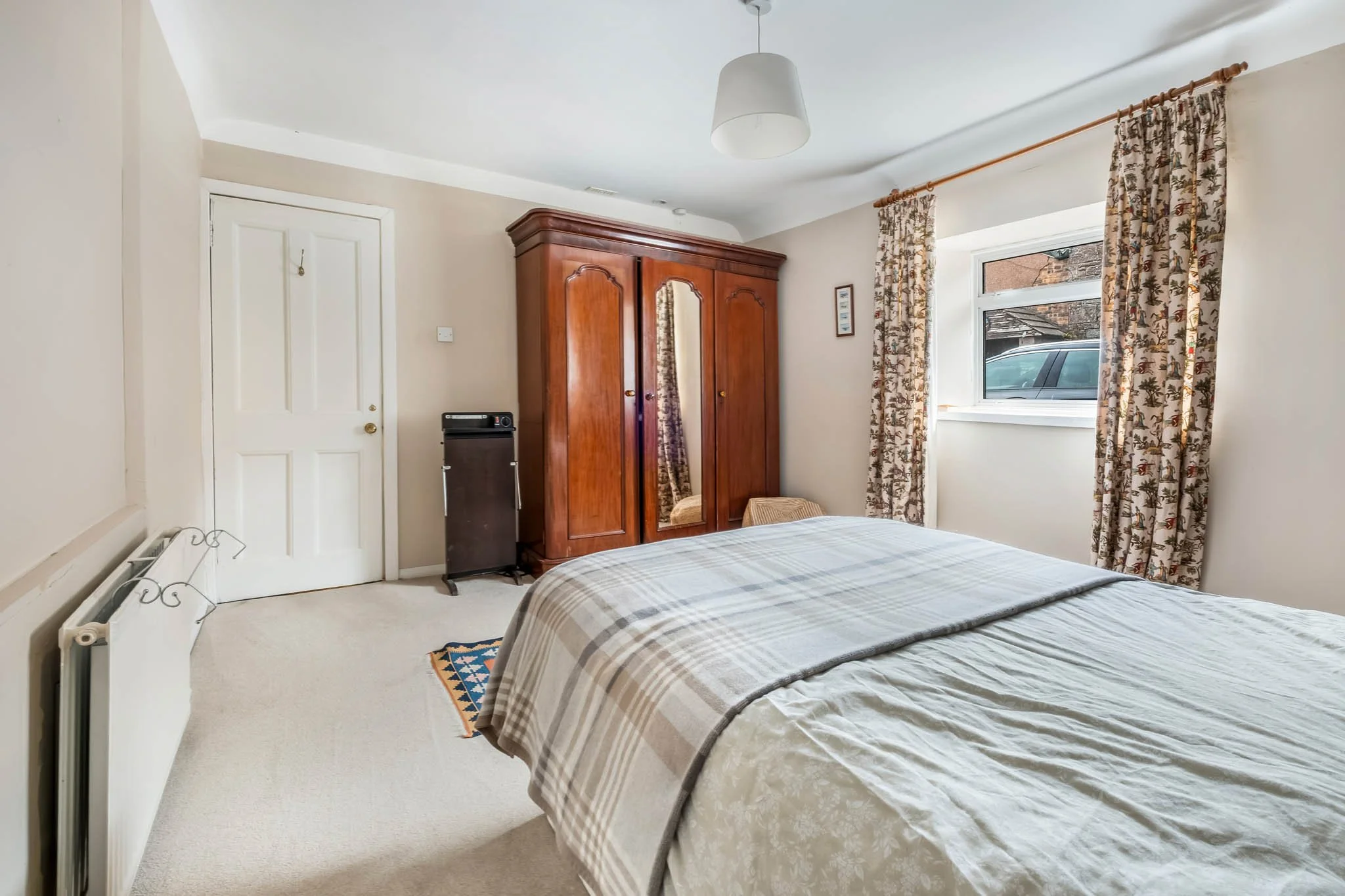 A bedroom with a bed covered in a light-colored plaid bedspread, a wooden wardrobe with a mirror, a window with patterned curtains, a small woven stool, and a white ceiling light fixture.