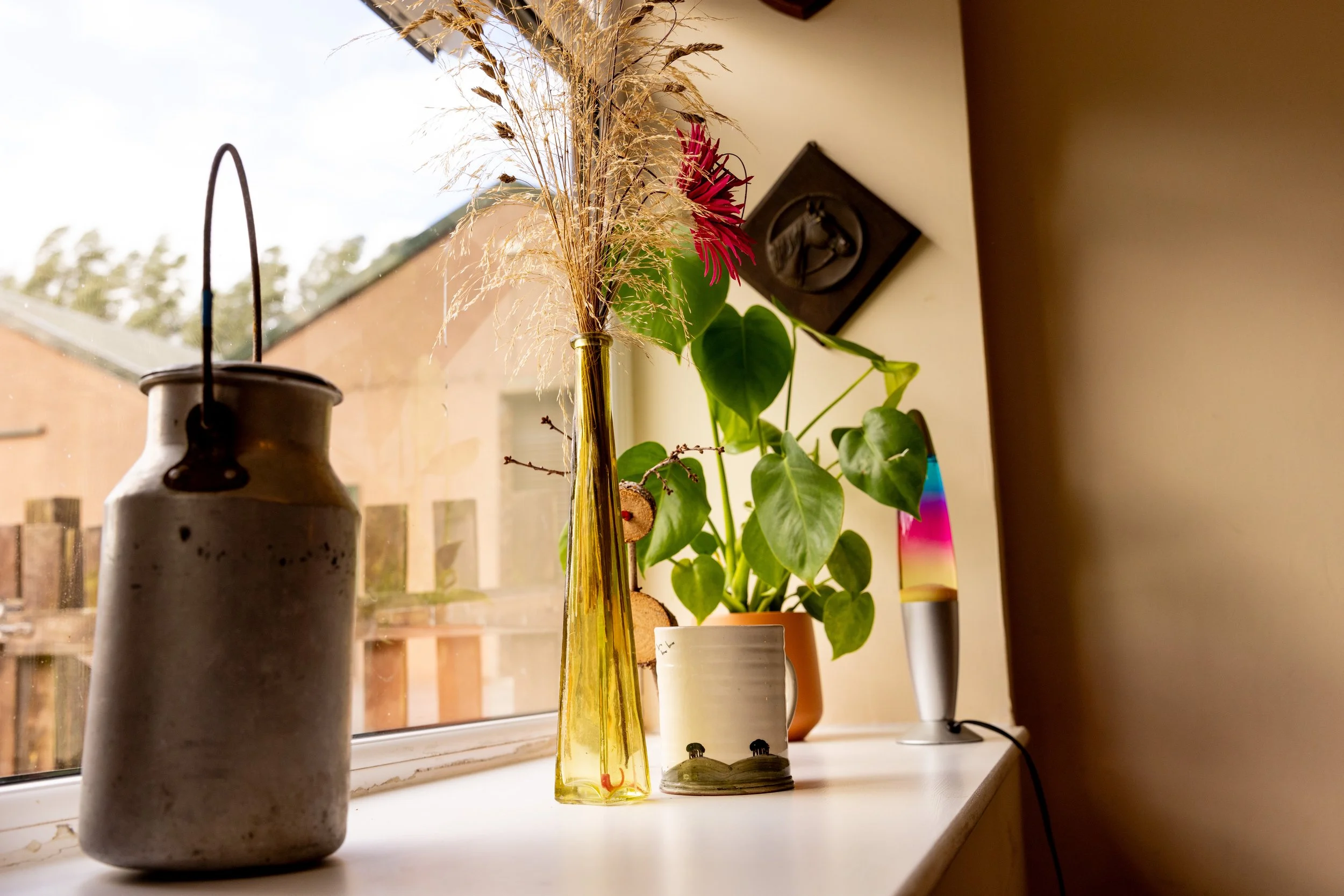 Decorative objects on a windowsill including a gray ceramic vase, a tall yellow glass vase with wheat and red flower, a potted plant with green heart-shaped leaves, a gray figurine, and a lava lamp, with a view of a backyard and wooden fence outside 